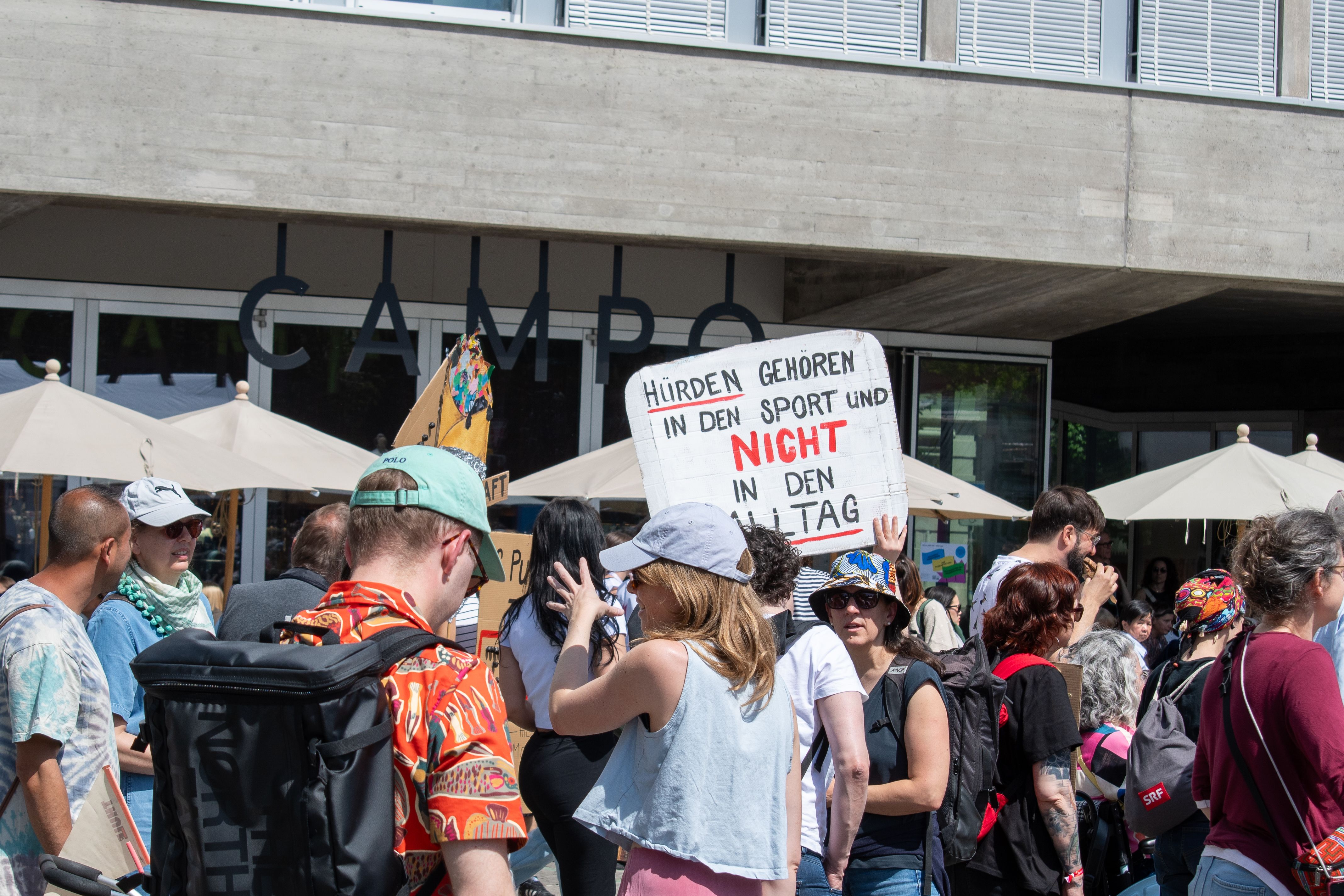 Draussen versammeln sich Menschen. Eine Person hält ein Schild mit der Aufschrift „Hürden gehören in den Sport und nicht in den Alltag“ hoch.