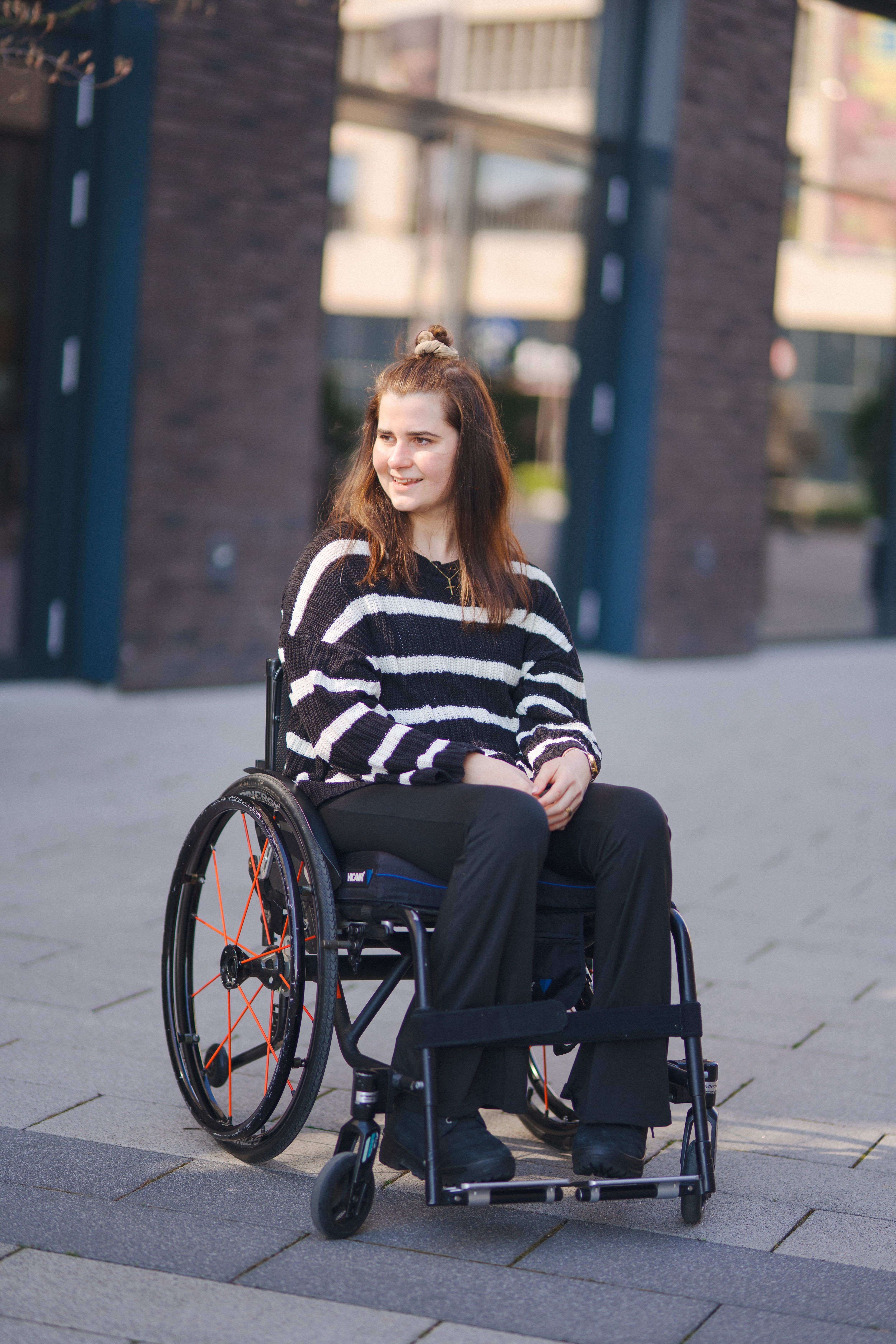 Mira Fischbacher in einem gestreiften Pullover sitzt draussen in einem Rollstuhl, lächelt und schaut zur Seite.