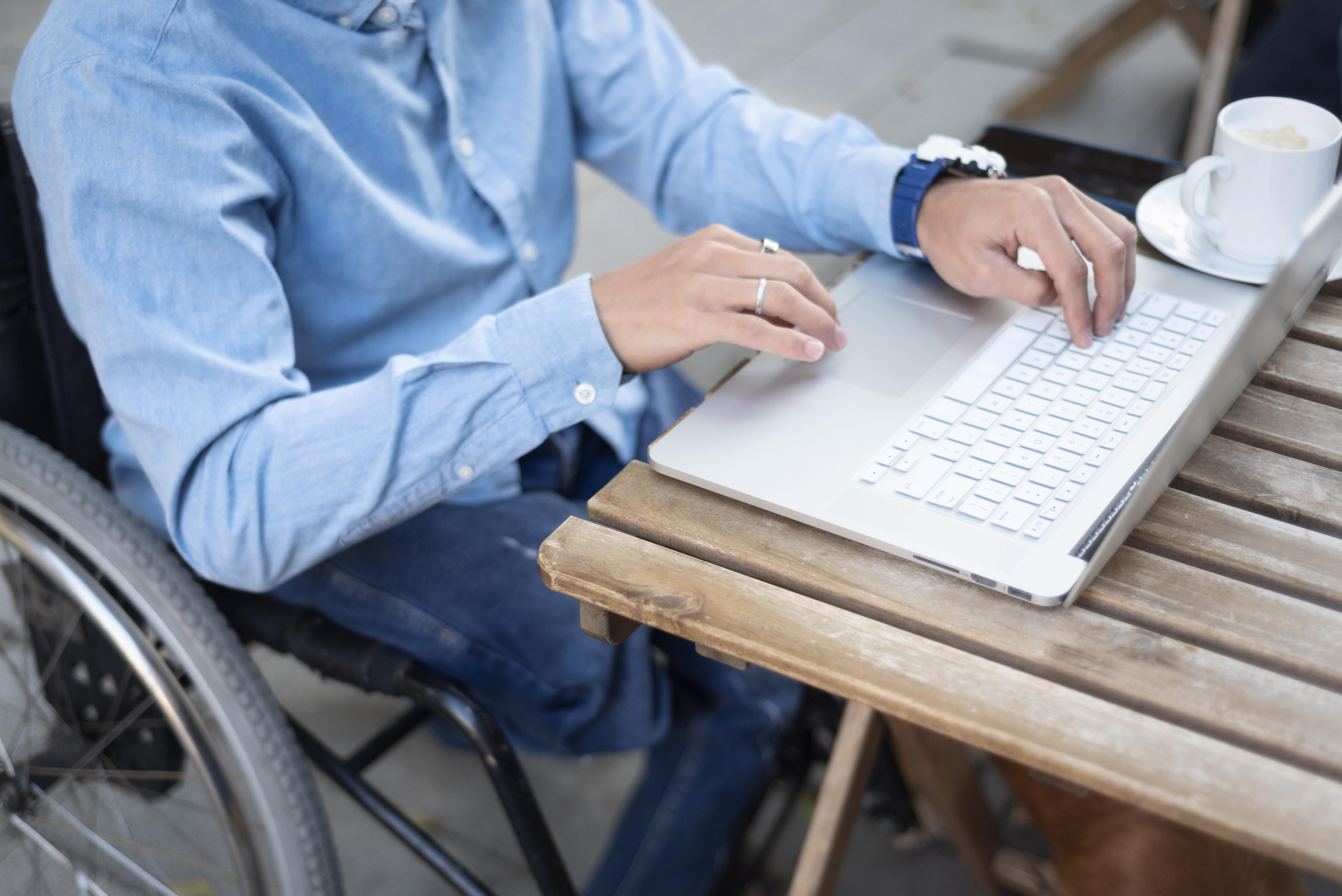 Eine Person im Rollstuhl, die an einem Holztisch an einem Laptop arbeitet, ein blaues Hemd trägt und neben sich eine Kaffeetasse stehen hat.