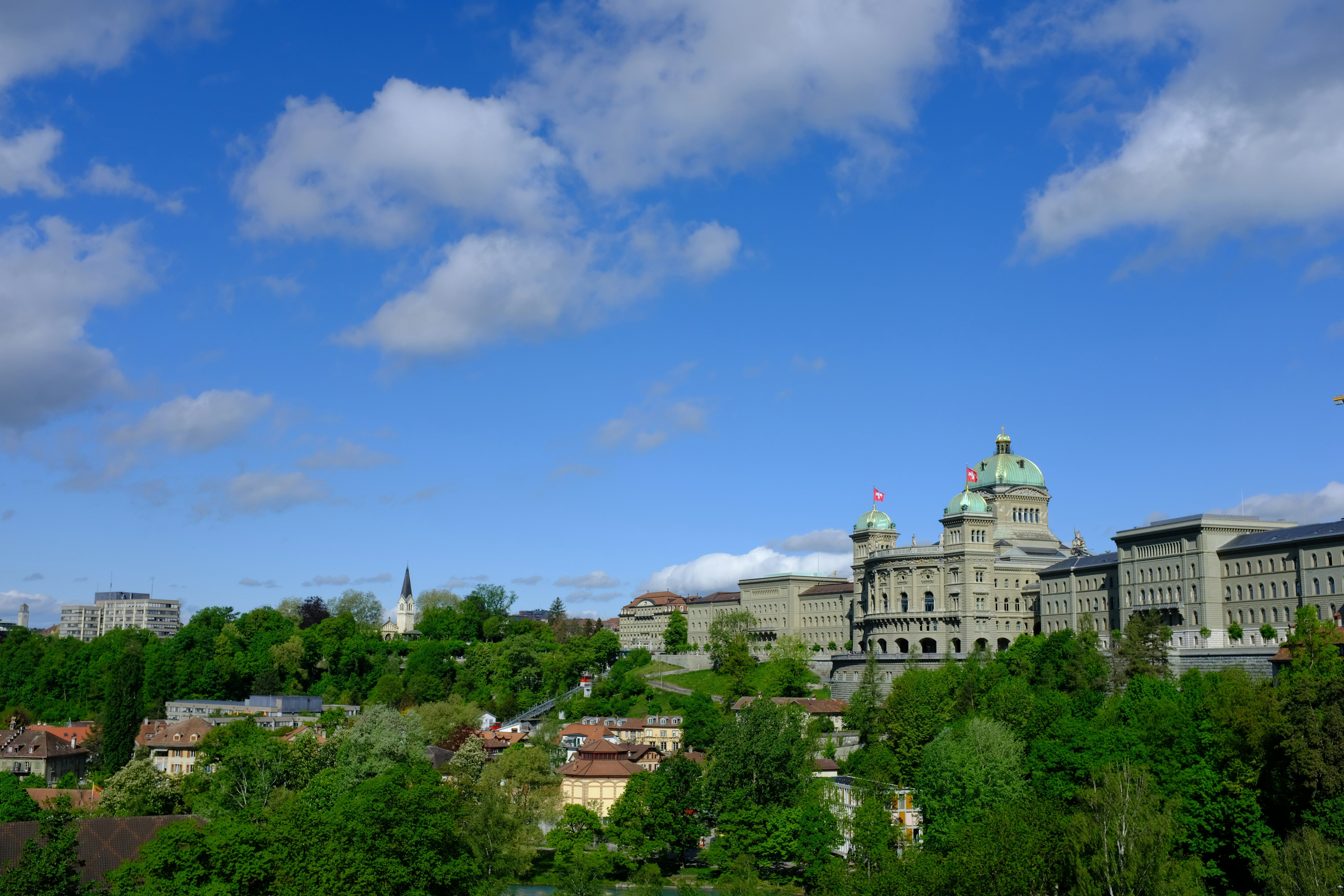 Grüne Landschaft mit Gebäuden und dem Bundeshaus unter einem blauen Himmel mit Wolken.