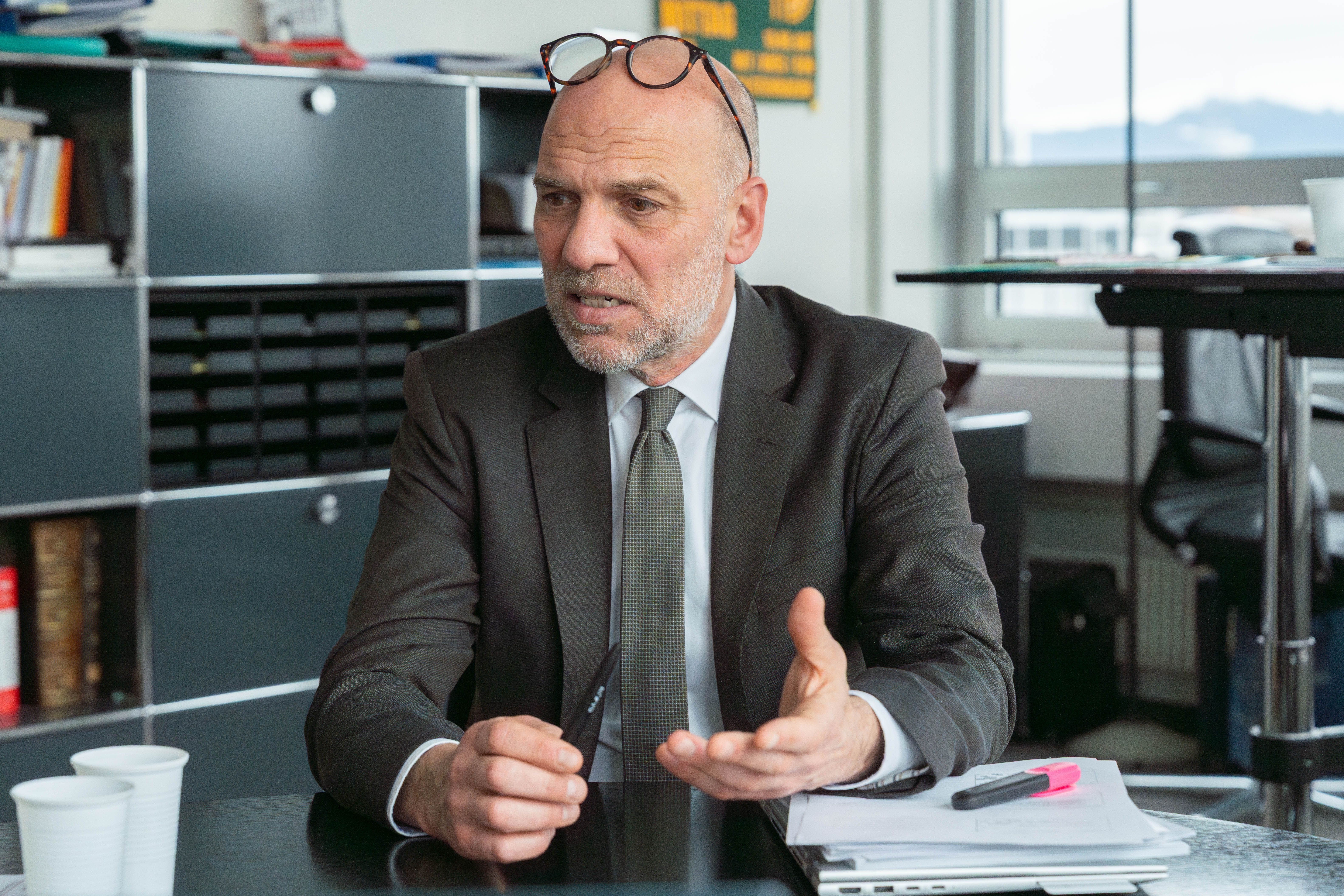 Florian Steinbacher hat eine Glatze, trägt einen Anzug und sitzt an einem Schreibtisch voller Papiere, spricht, hält einen Stift in der Hand und trägt eine Brille auf dem Kopf. Im Hintergrund sind Regale und ein Fenster zu sehen.
