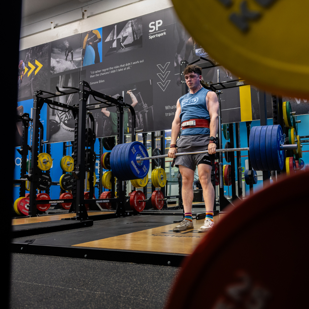 A young man in a blue tank top and red weight belt lifts a heavy barbell with blue plates in a modern gym.