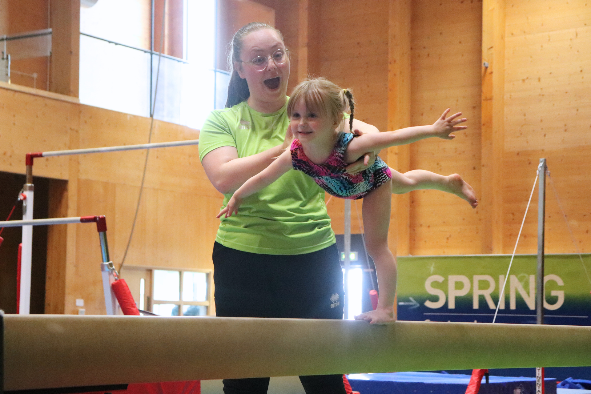 Teacher helps little girl on gymnastics balance beam to balance on one leg