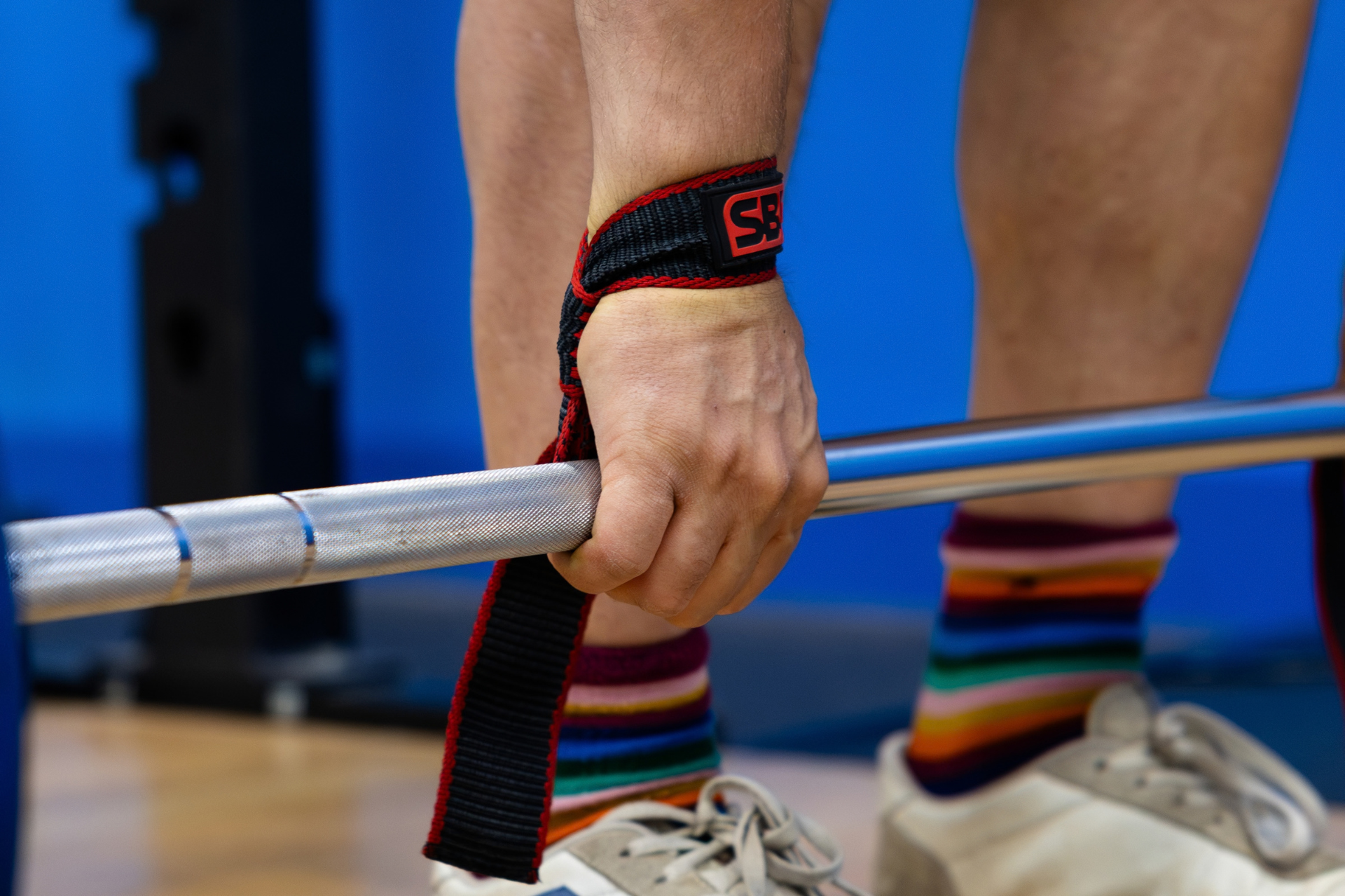 A close-up of a weightlifter gripping a barbell with a black and red wrist strap, with colorful striped socks visible in the background.