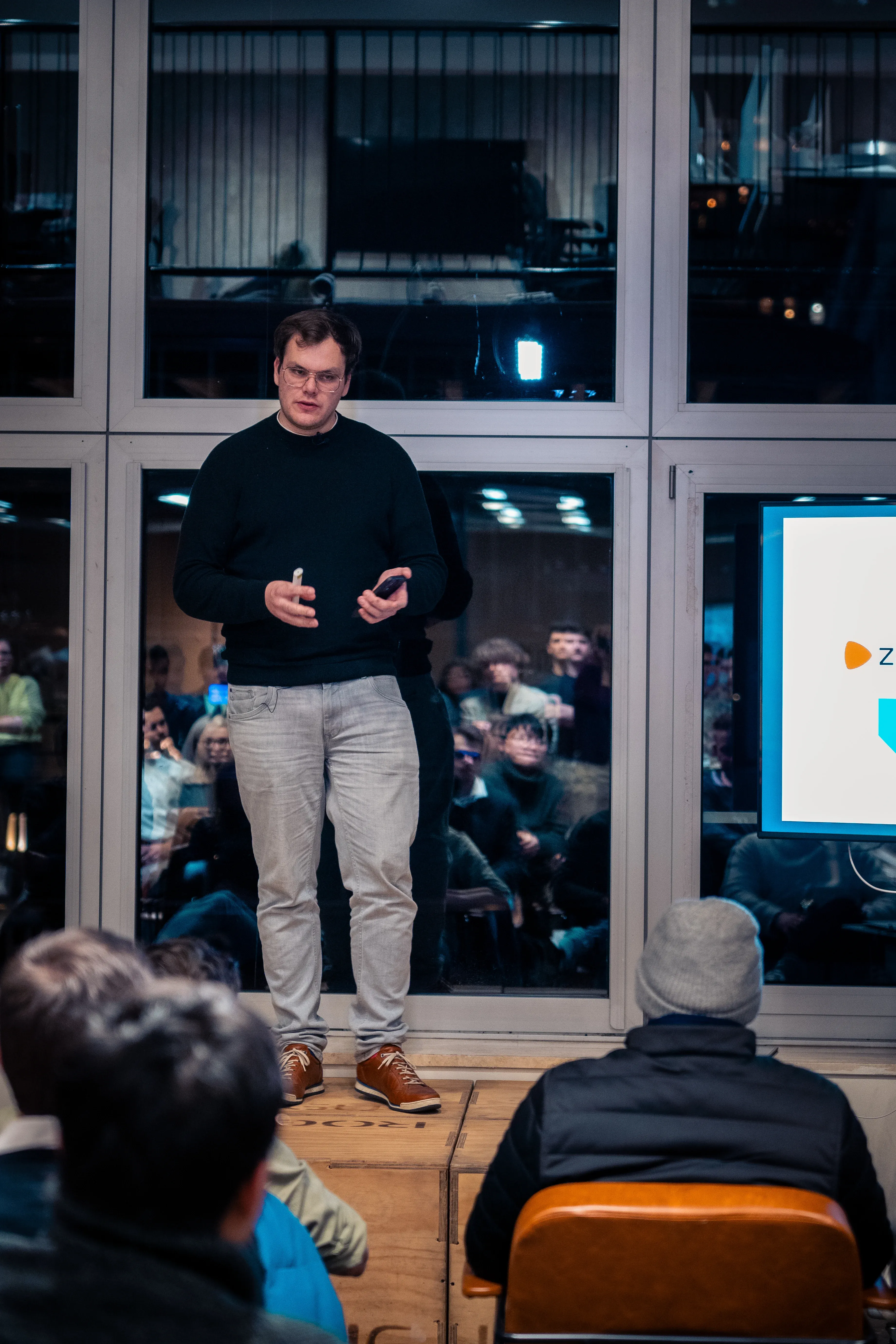 A person in a black sweater and light pants speaks to an audience, standing on a platform in front of a large window and a presentation screen.