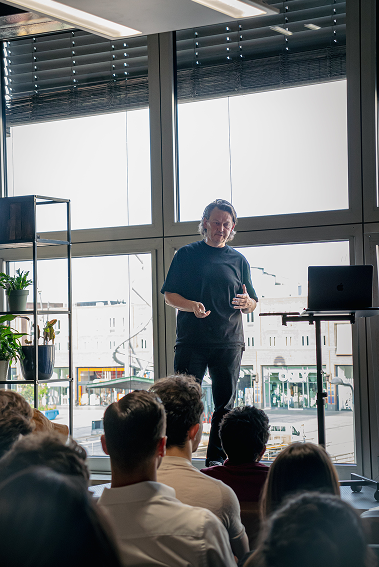 A person in a black shirt presents at the design meetup in a bright room with large windows, a laptop, and a shelf with plants.