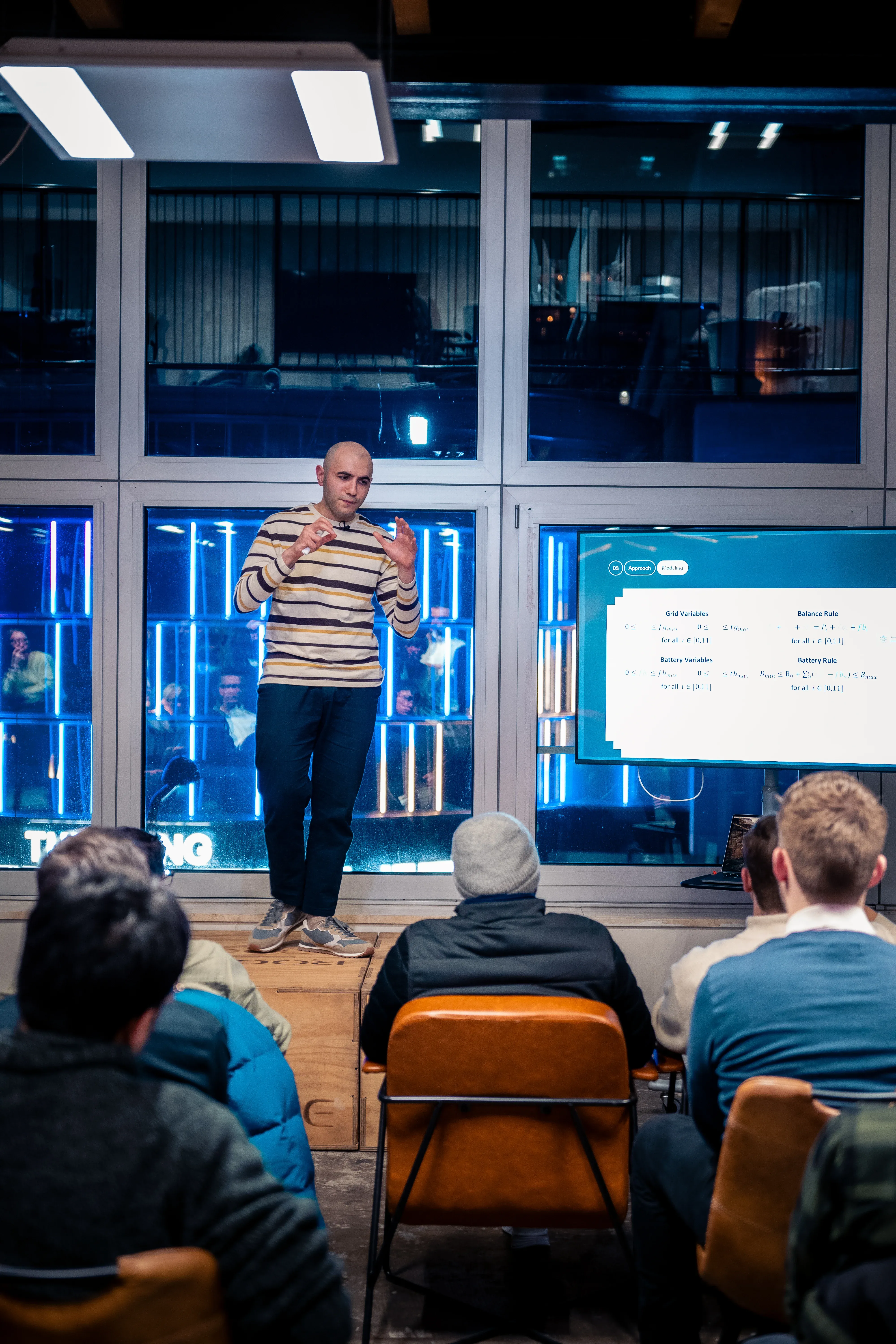 A man stands on a box, giving a presentation to an audience. A screen displays information beside him.