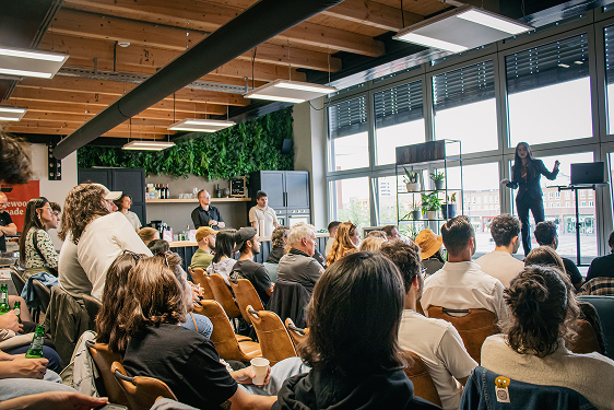 A speaker addresses a seated audience at the design meetup in a room with large windows and a green wall.