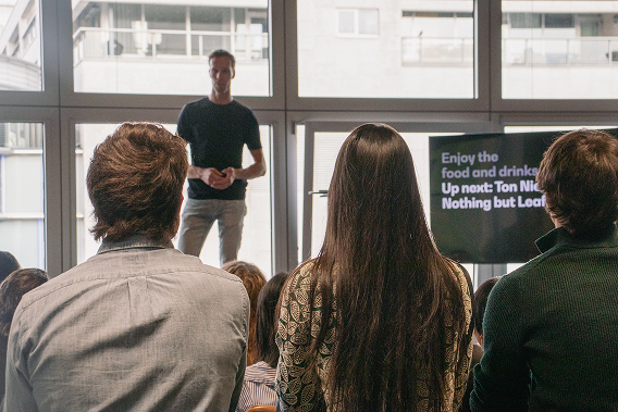 A speaker stands in front of an audience at the design meetup, with a screen displaying event information in the background.