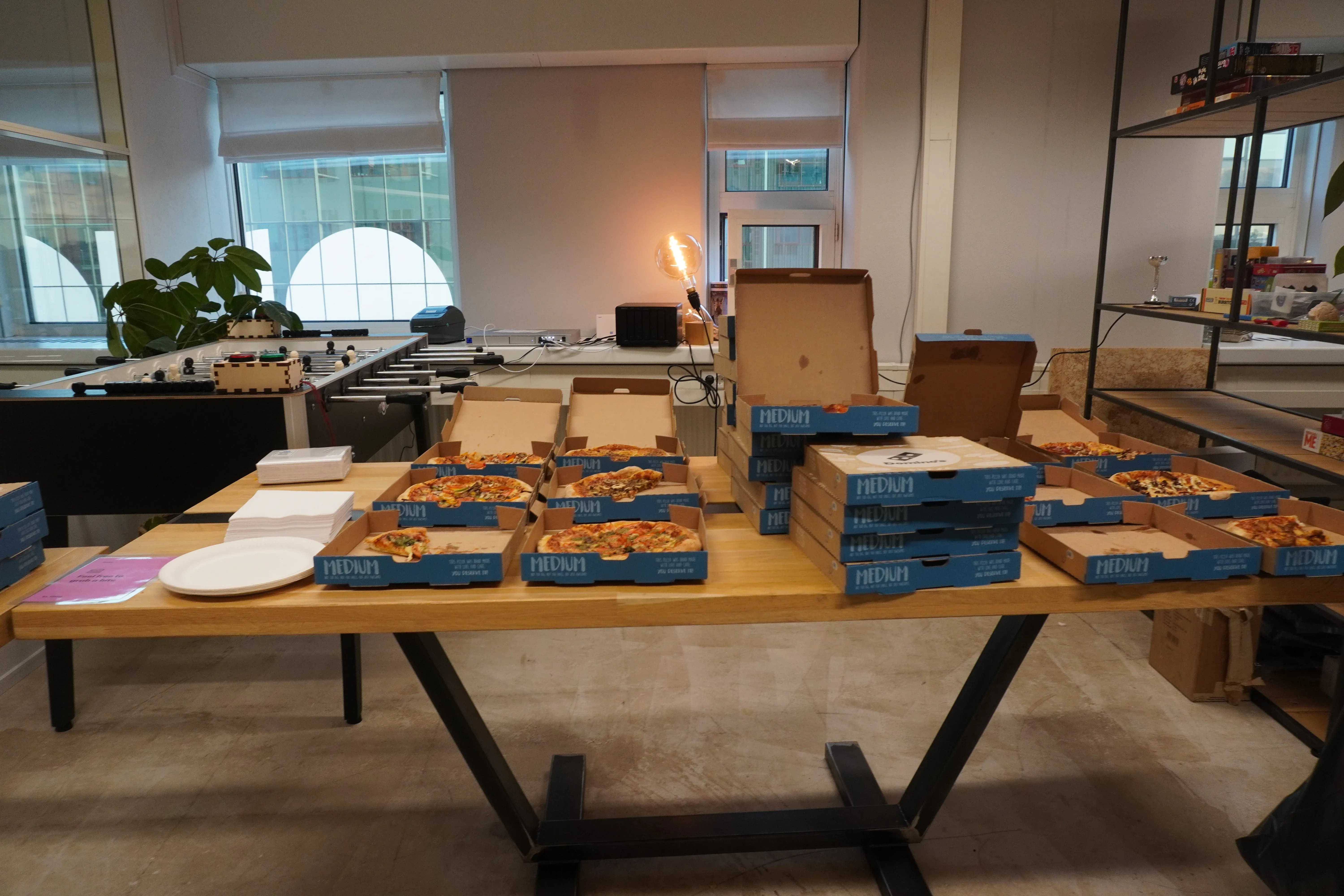A table with multiple open pizza boxes, some empty plates, and a foosball table in the background in an office setting.