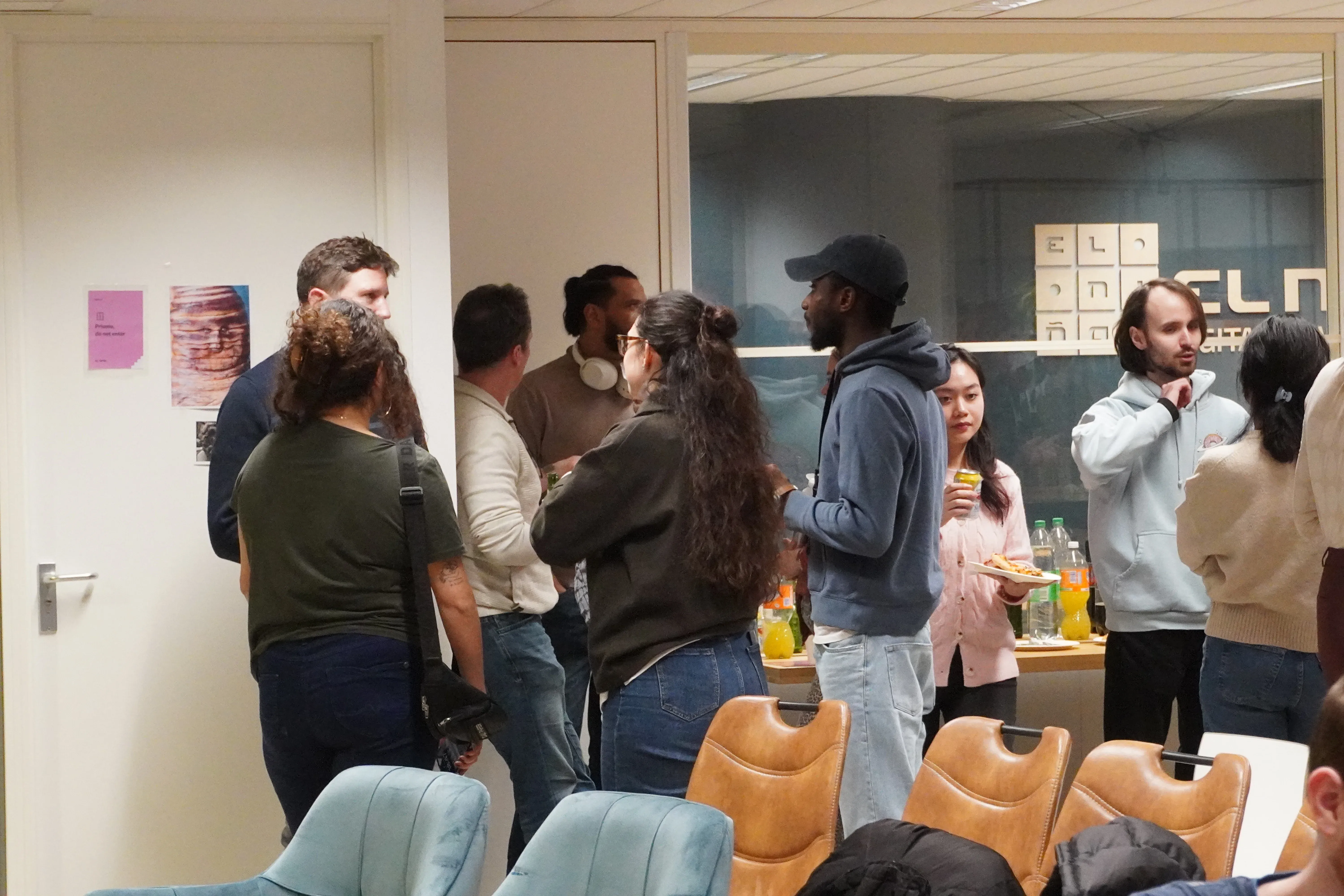 A group of people socializing in an office, standing near a table with drinks and snacks, some holding cups and plates.