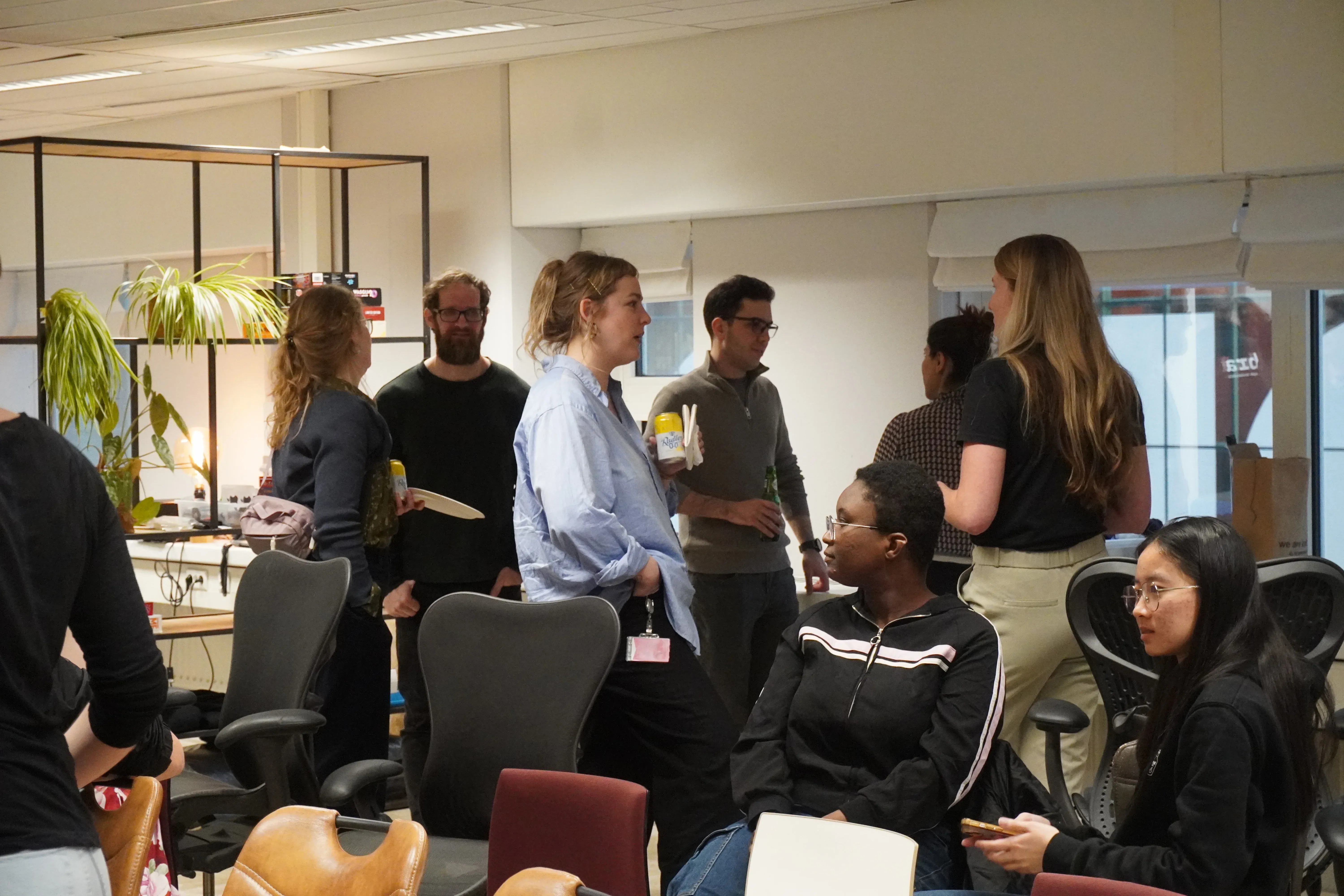 A group of people in an office setting, standing and sitting, engaged in conversation. Office furniture and plants are visible in the background.