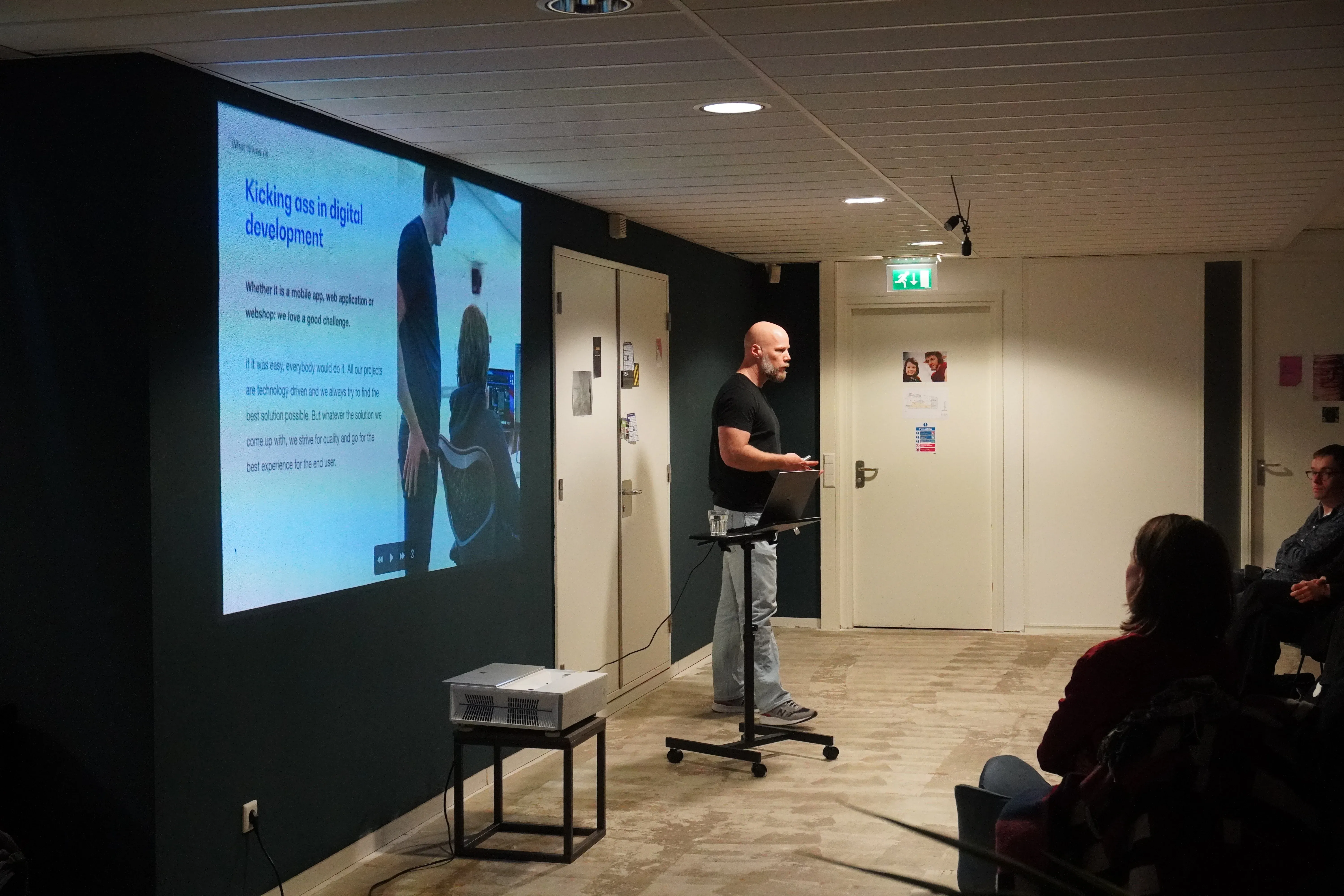 A person giving a presentation on digital development in a dimly lit room, with a slide displayed on a large screen. Audience members seated.