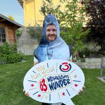 Person in Fischkostüm hält ein großes Schild mit der Aufschrift "Ö3 Weihnachtswunder" auf einer Wiese vor einem Haus.