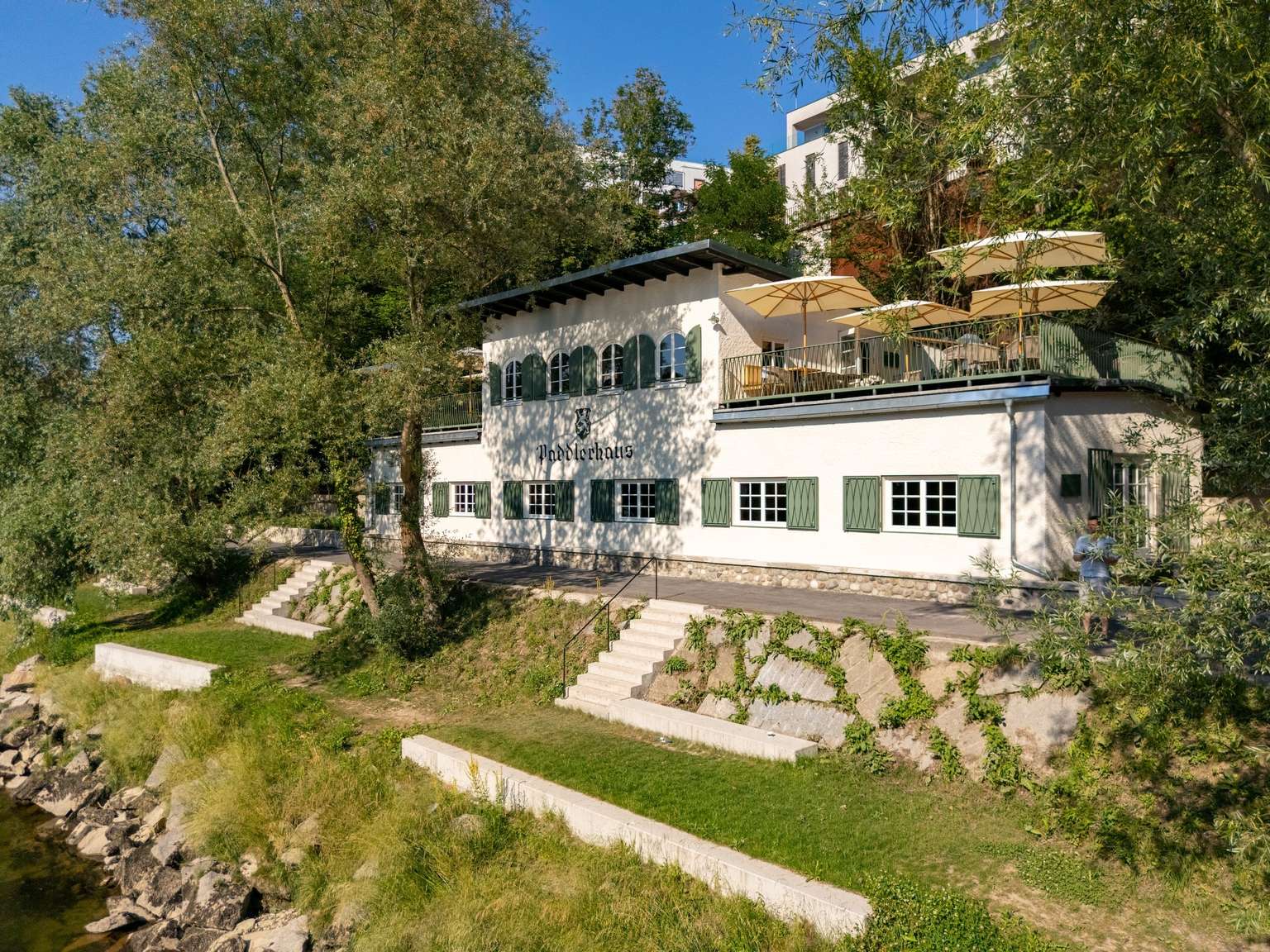 Das revitalisierte Paddlerhaus am Ennsufer in Steyr mit grünen Fensterläden und Sonnenschirmen auf der Terrasse
