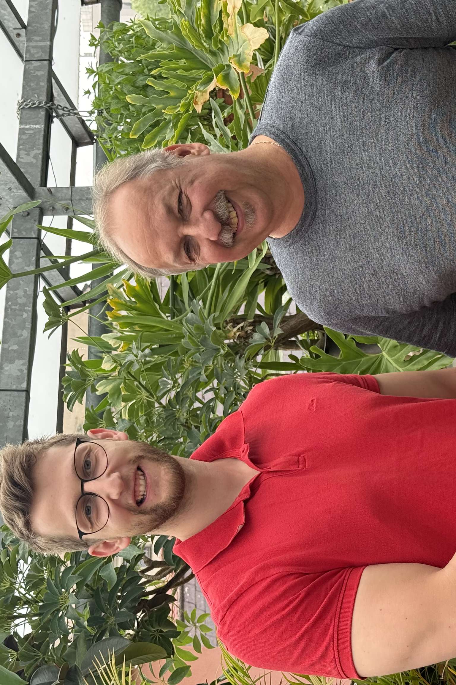 Zwei Männer stehen in einem grünen Gewächshaus und schütteln sich die Hände. Der eine trägt ein rotes Shirt, der andere ein graues.