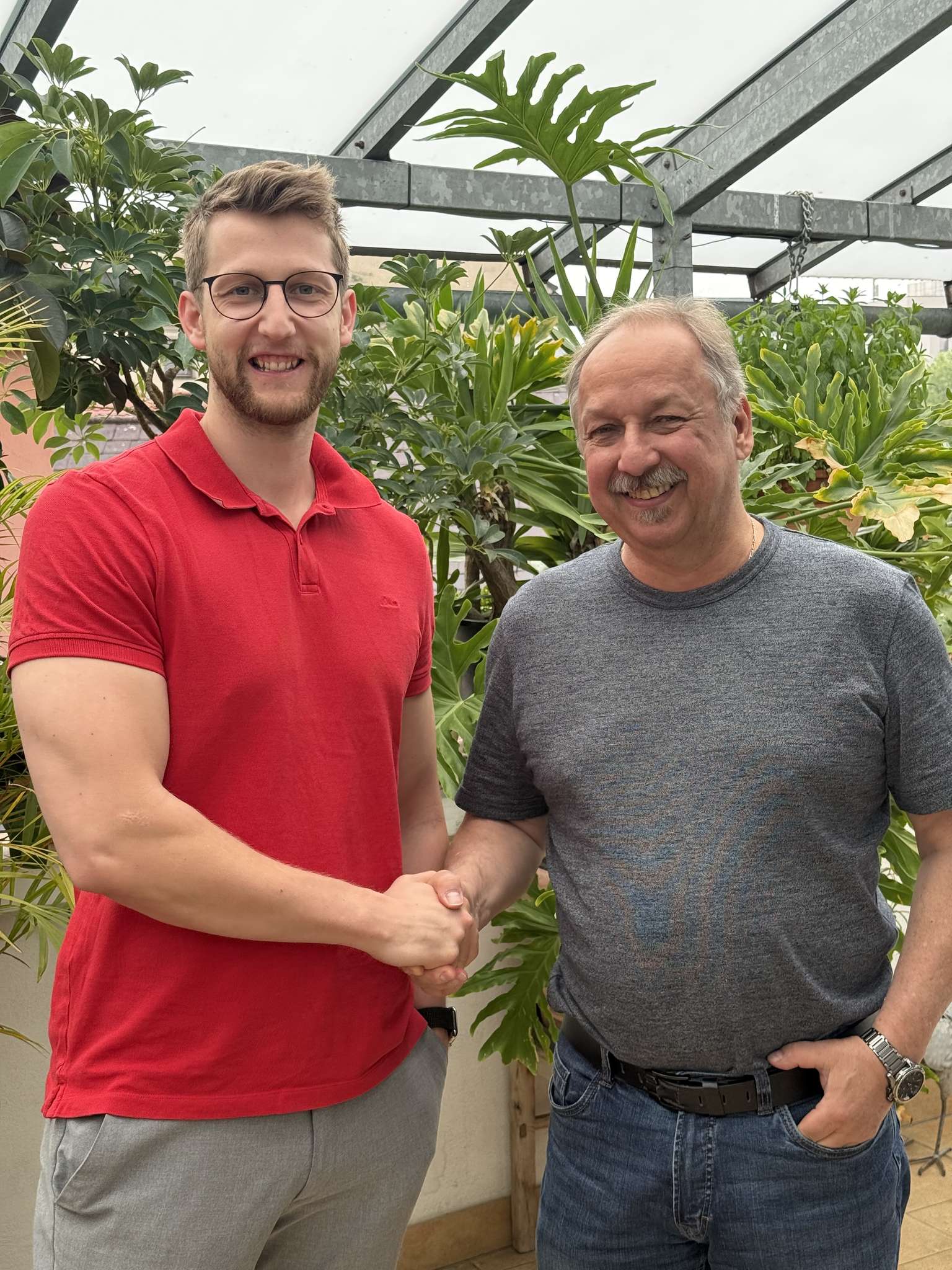 Zwei Männer stehen in einem grünen Gewächshaus und schütteln sich die Hände. Der eine trägt ein rotes Shirt, der andere ein graues.