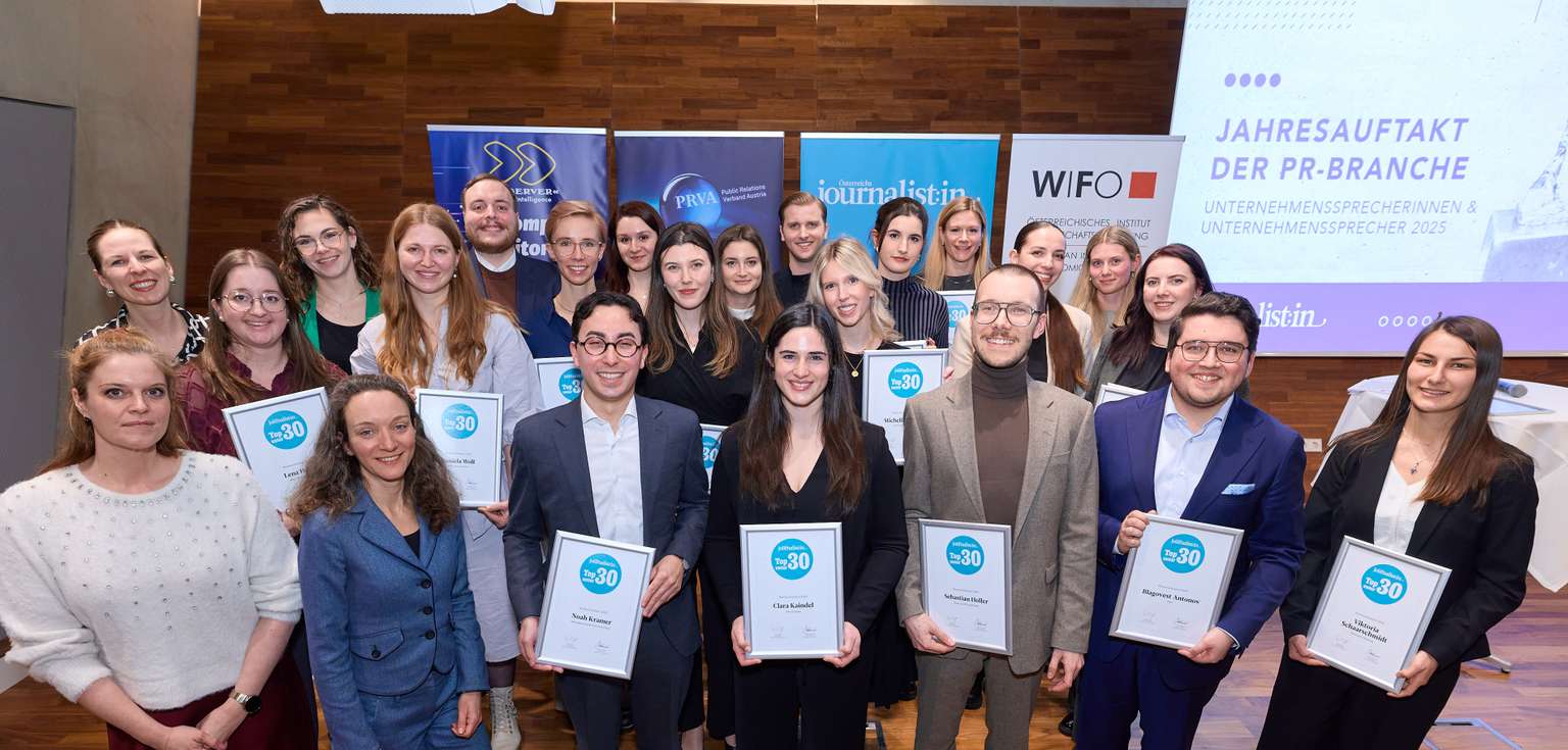 Group photo of smiling people holding certificates, standing in front of banners and a screen at an event in a wood-paneled room.