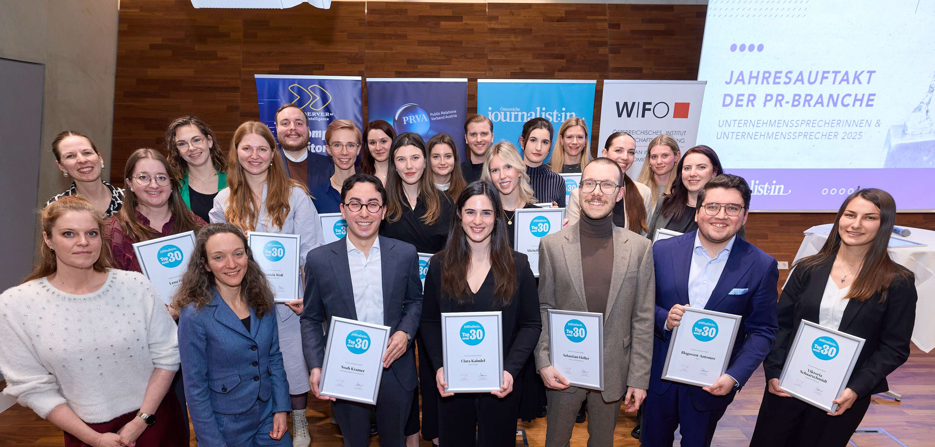 Group photo of smiling people holding certificates, standing in front of banners and a screen at an event in a wood-paneled room.