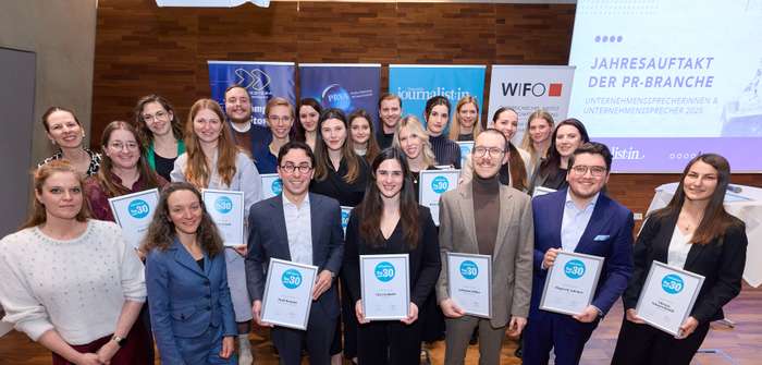 Group photo of smiling people holding certificates, standing in front of banners and a screen at an event in a wood-paneled room.