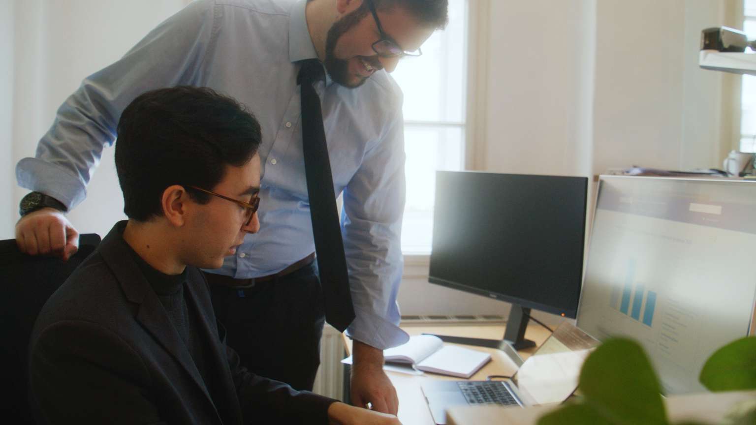 Zwei männliche Personen bei einer Besprechung vor einem Schreibtisch in einem hellen Raum mit grünen Pflanzen