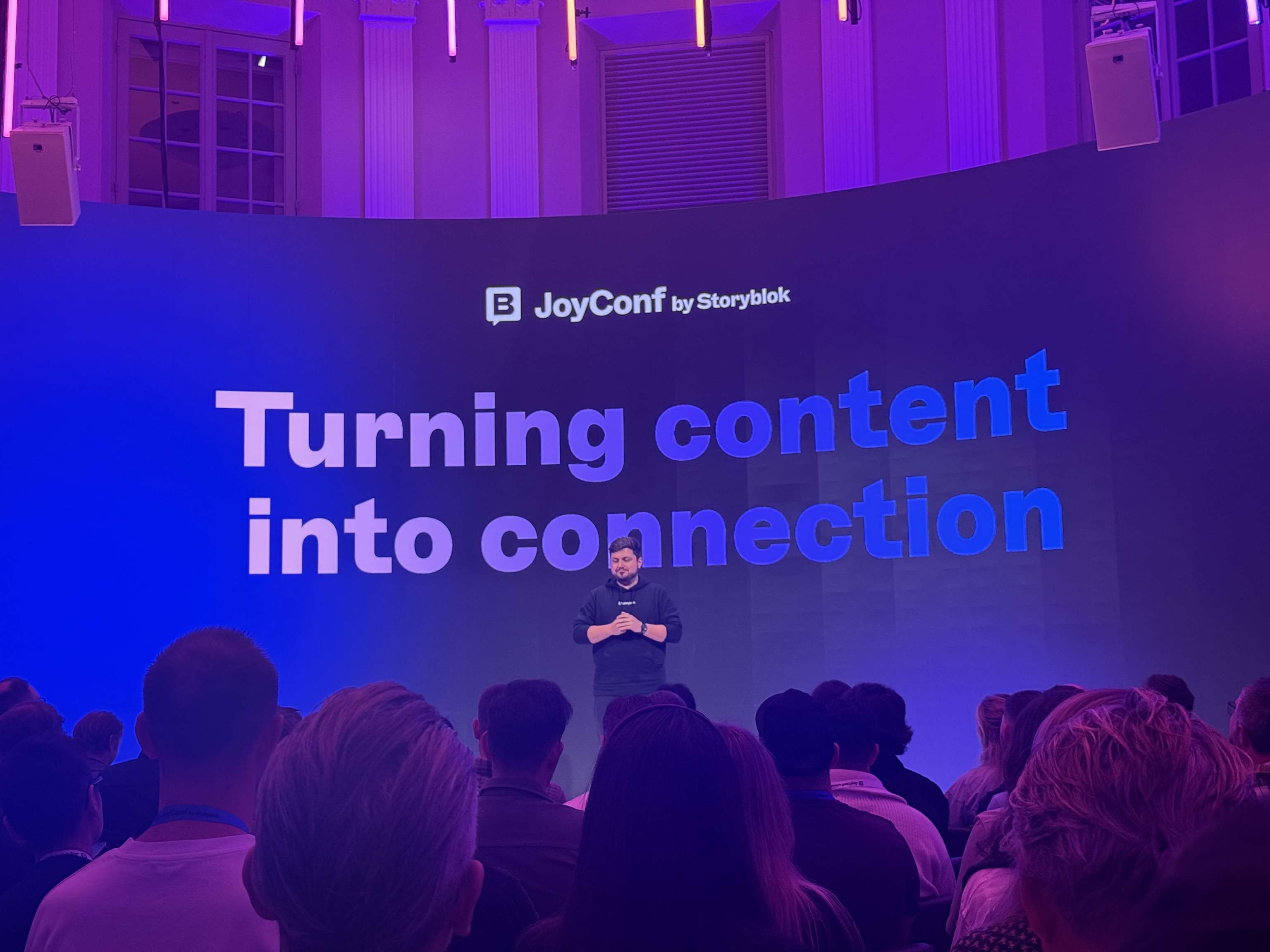 A speaker stands on stage at JoyConf by Storyblok, addressing an audience with the message "Turning content into connection" on the screen.