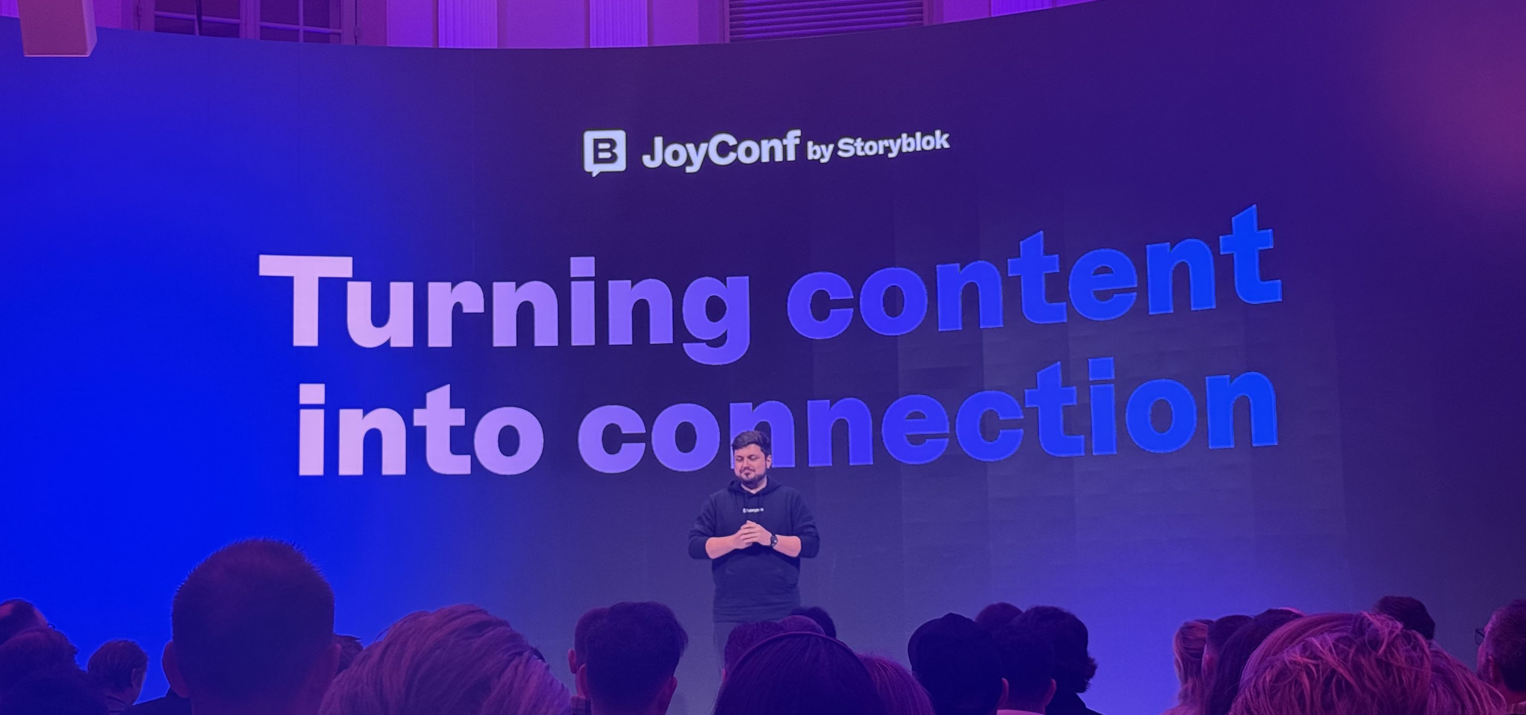 A speaker stands on stage at JoyConf by Storyblok, addressing an audience with the message "Turning content into connection" on the screen.