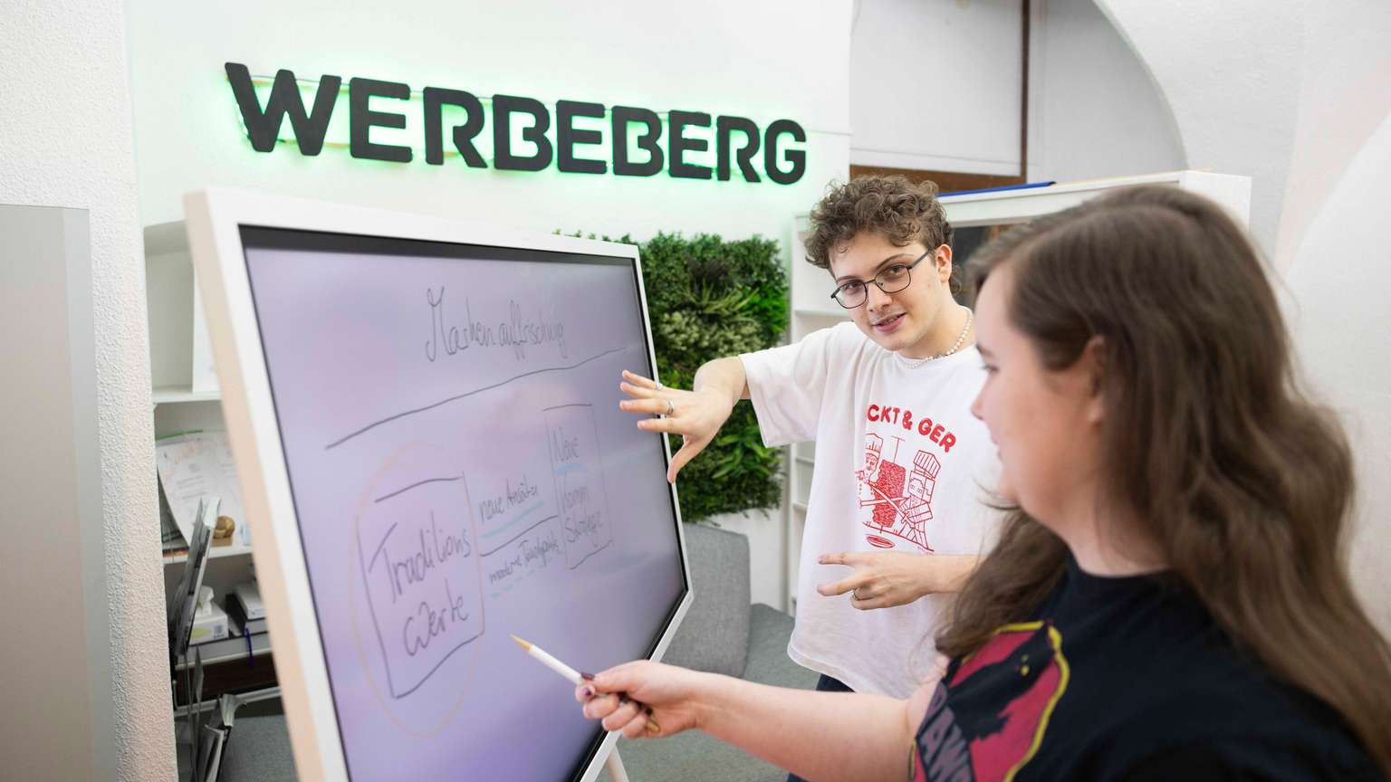 Zwei junge Menschen stehen vor einem Whiteboard und diskutieren.