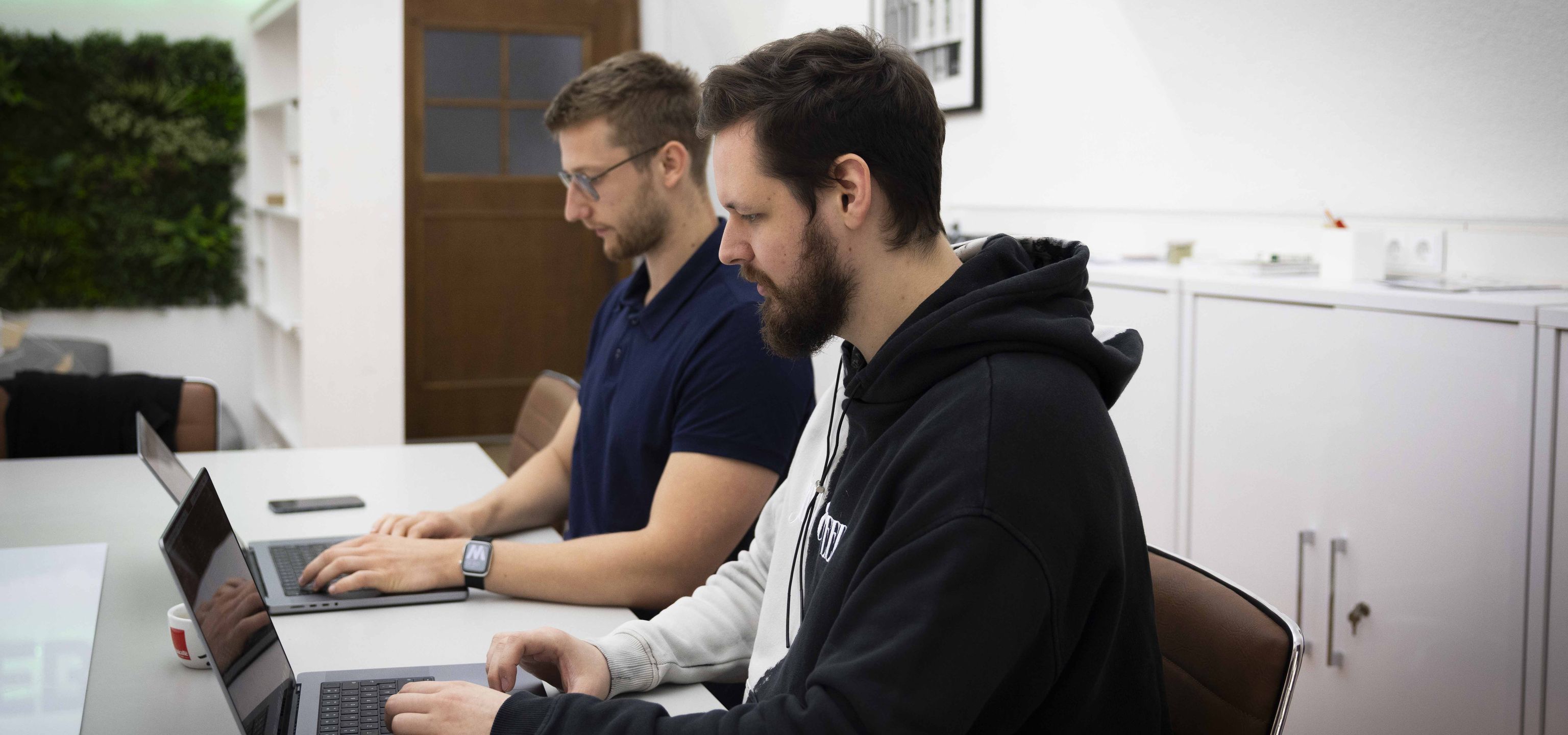 Zwei junge Männer sitzen nebeneinander und arbeiten an ihren Laptops
