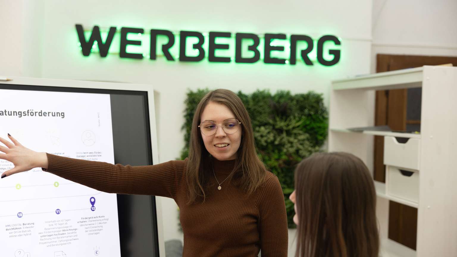 Zwei junge Frauen reden und zeigen auf ein digitales Flipboard.