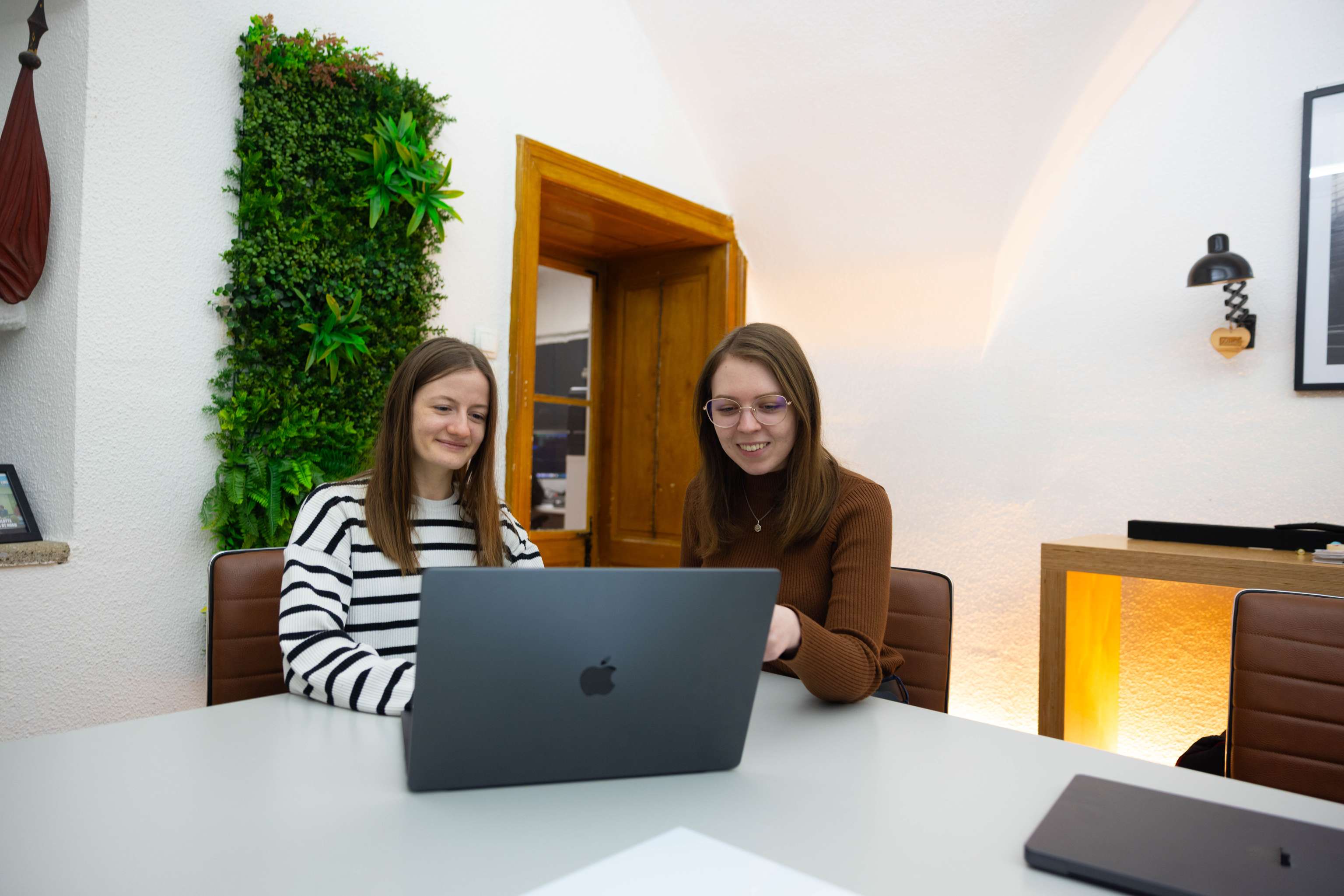 Zwei Frauen, die in Büroumgebung hinter Laptop sitzen und lächelnd auf den Bildschirm sehen.