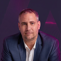 Mann im dunkelblauen Anzug vor einem geometrischen, violetten Hintergrund. Er stützt sich mit den Armen in entspannter Pose ab.