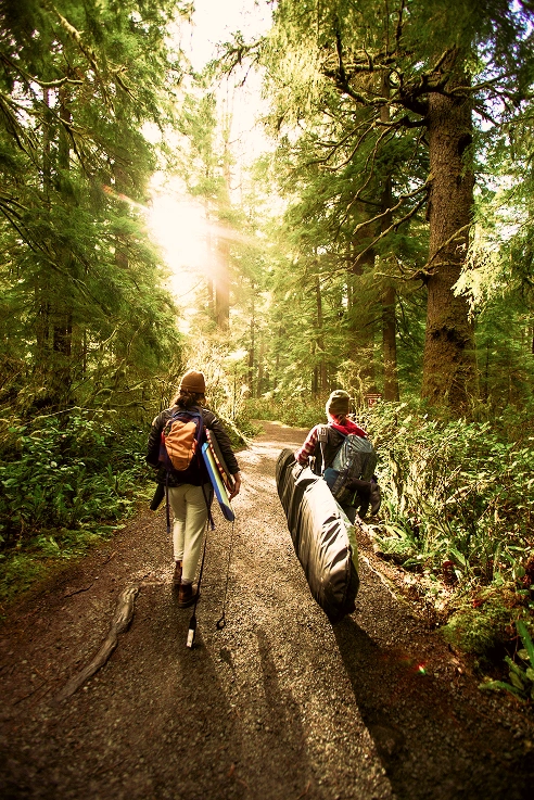 Zwei Menschen gehen mit Surfbrettern auf einem Waldweg. Sonnenlicht fällt durch das dichte grüne Laub und schafft eine ruhige Atmosphäre.