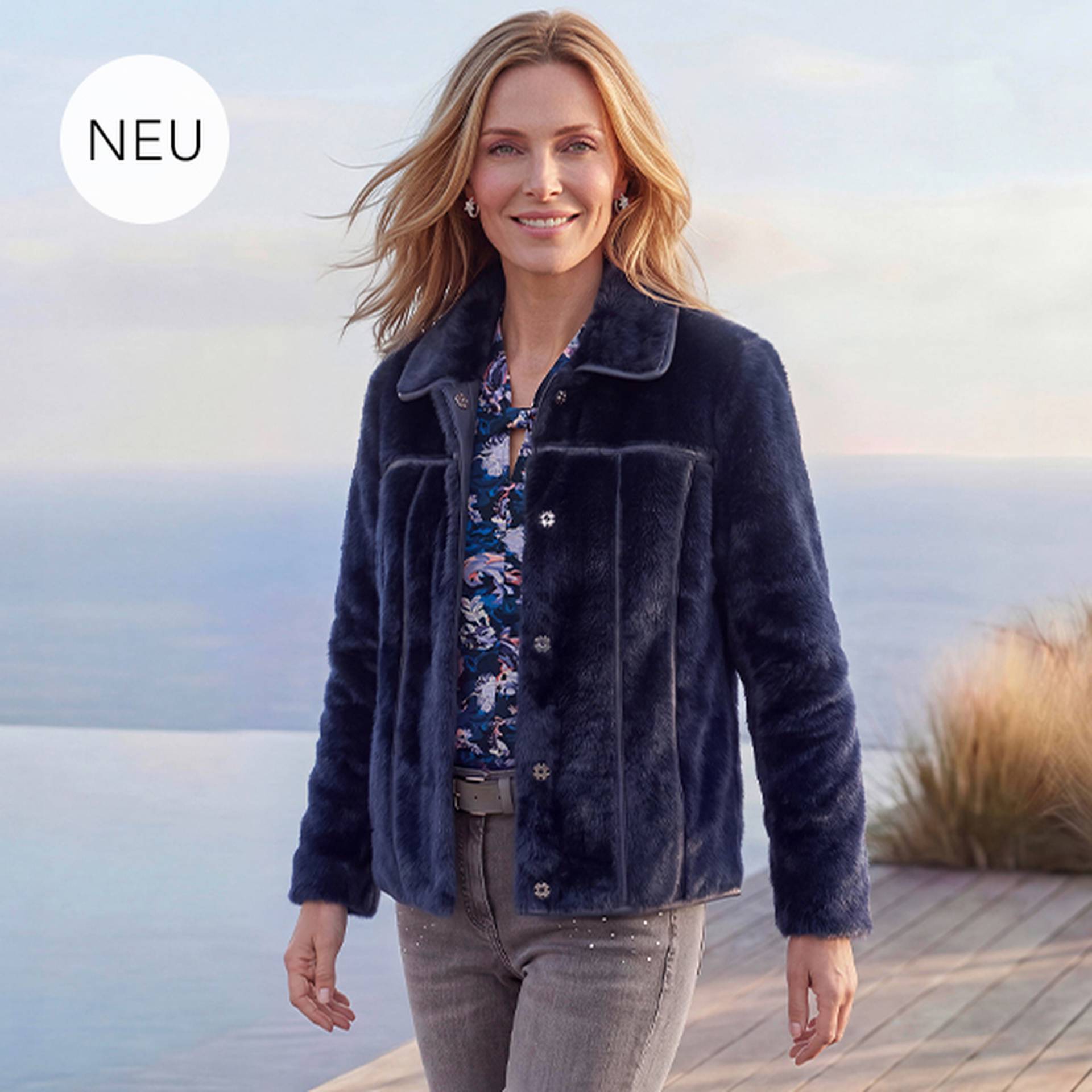 Eine Frau Eine Frau, die eine marineblaue Wendejacke mit Lederoptik und Kunstfell über einer geblümten Bluse trägt und im Freien ein angenehmes Herbstwetter genießt.einer hellrosa Bluse und einer gemusterten Trendhose sitzt auf einer Treppe und lächelt im Sonnenlicht.