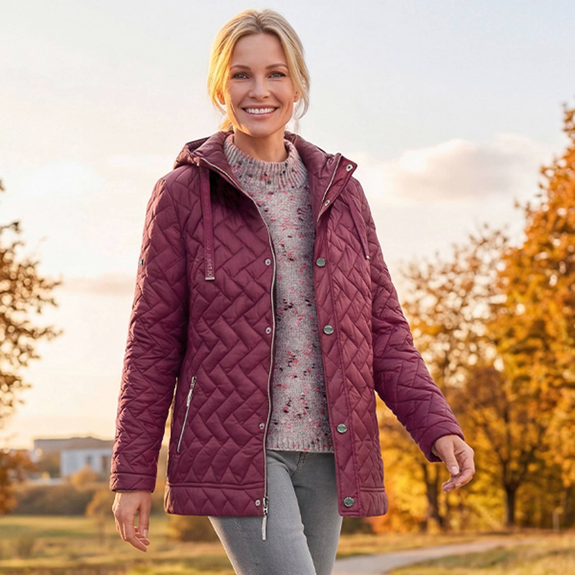 Eine Frau in einer burgunderroten Steppjacke und einer grauen Hose geht im Freien spazieren, im Hintergrund sind herbstliche Bäume zu sehen.
