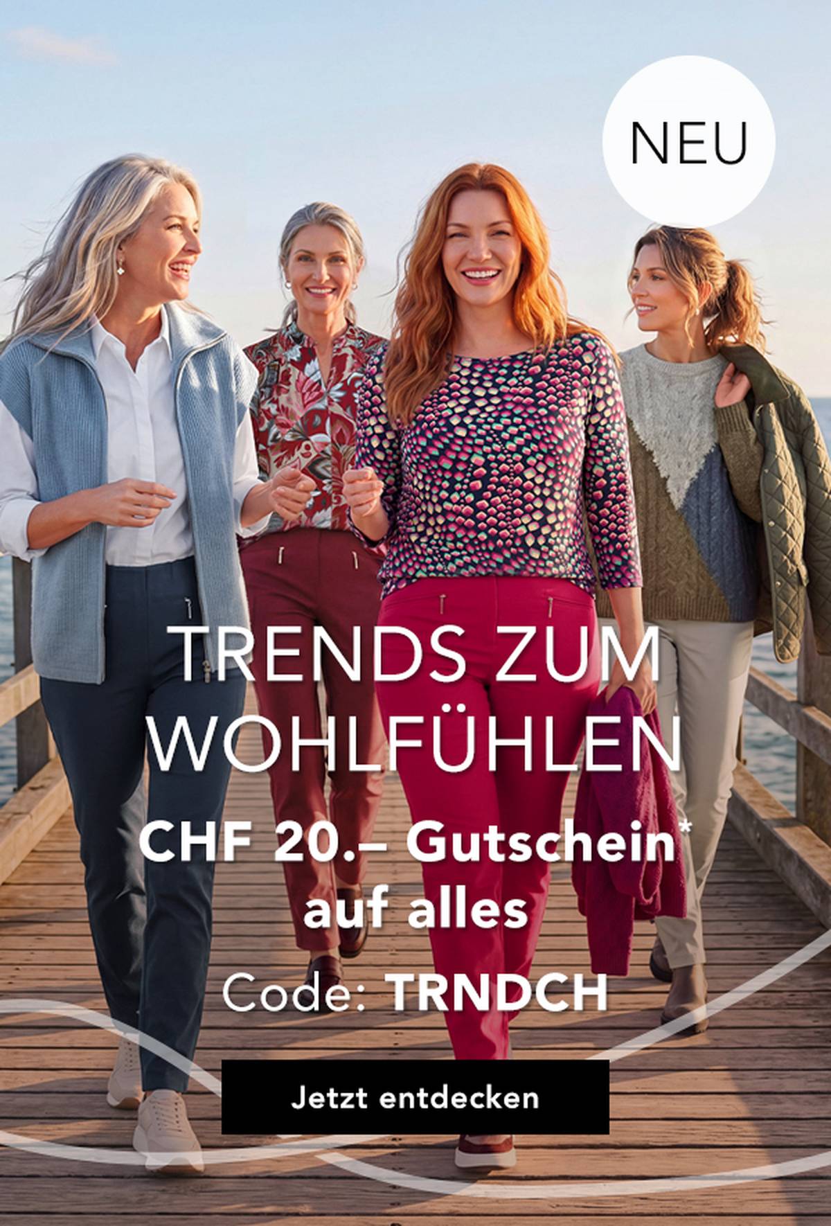 Vier Frauen in stilvollen, farbenfrohen Outfits spazieren auf einem Steg am Meer entlang, lächeln und genießen einen sonnigen Tag.