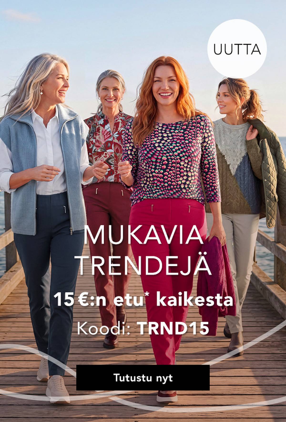 Neljä naista tyylikkäissä, värikkäissä asuissa kävelee laituria pitkin meren rannalla, hymyillen ja nauttien aurinkoisesta päivästä.