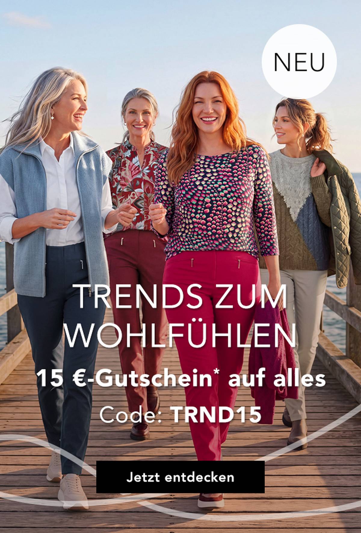 Vier Frauen in stilvollen, farbenfrohen Outfits spazieren auf einem Steg am Meer entlang, lächeln und genießen einen sonnigen Tag.