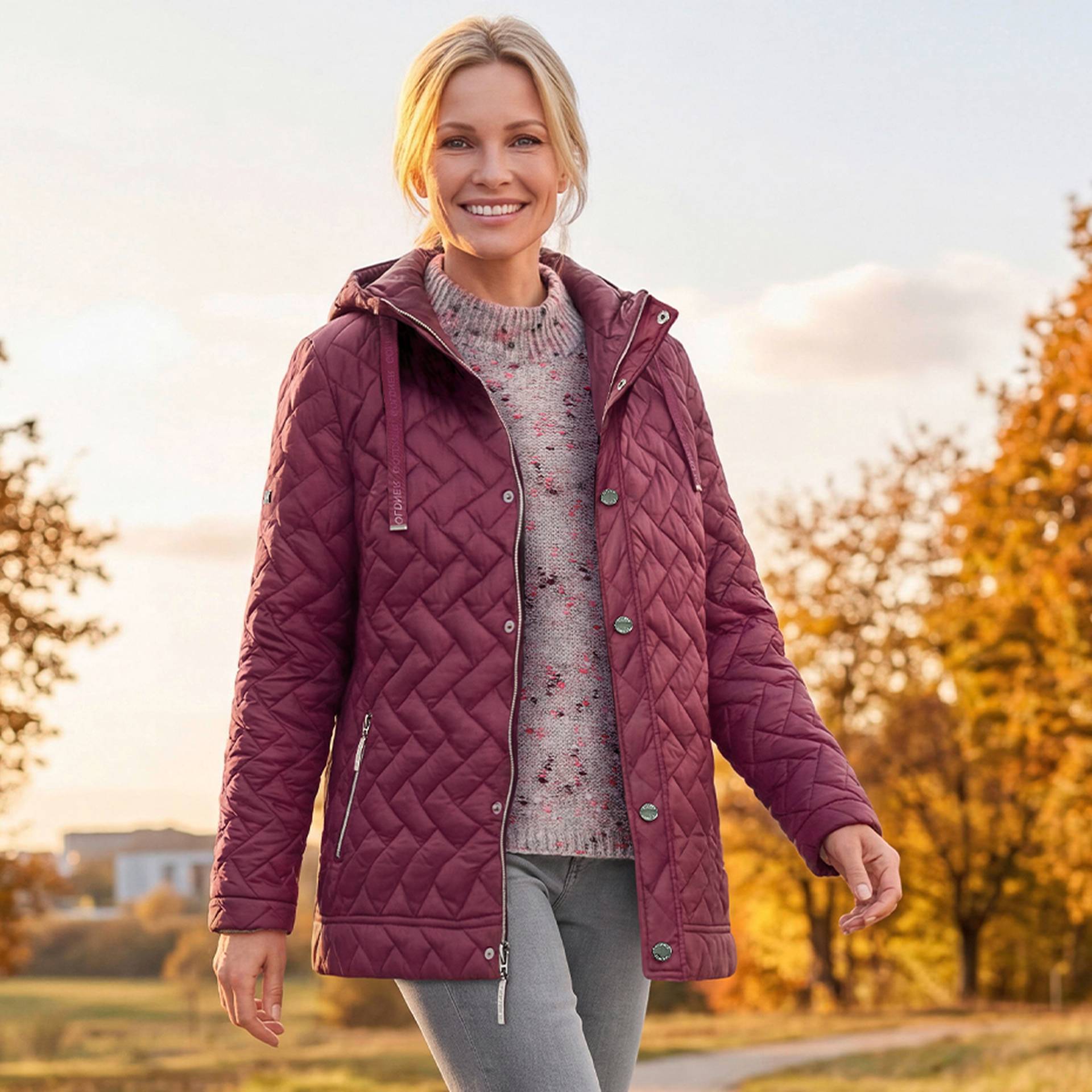 Eine Frau in einer burgunderroten Steppjacke und einer grauen Hose geht im Freien spazieren, im Hintergrund sind herbstliche Bäume zu sehen.