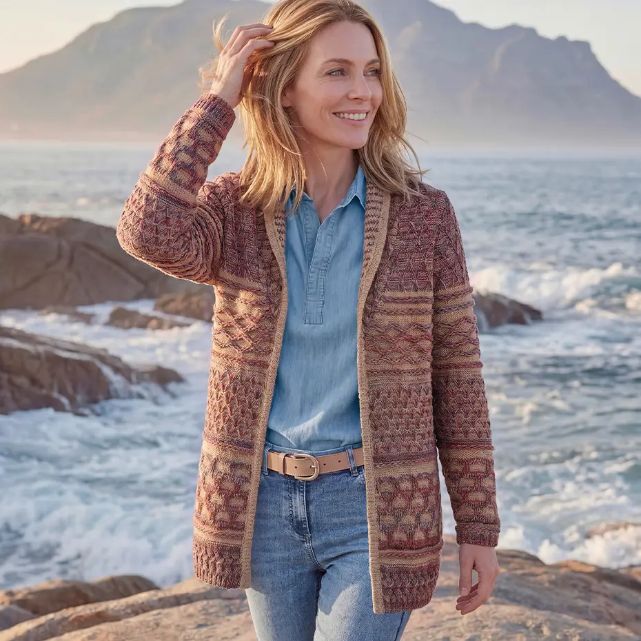 Frau in elegantem Strickcardigan und Jeans steht lächelnd am Meer.