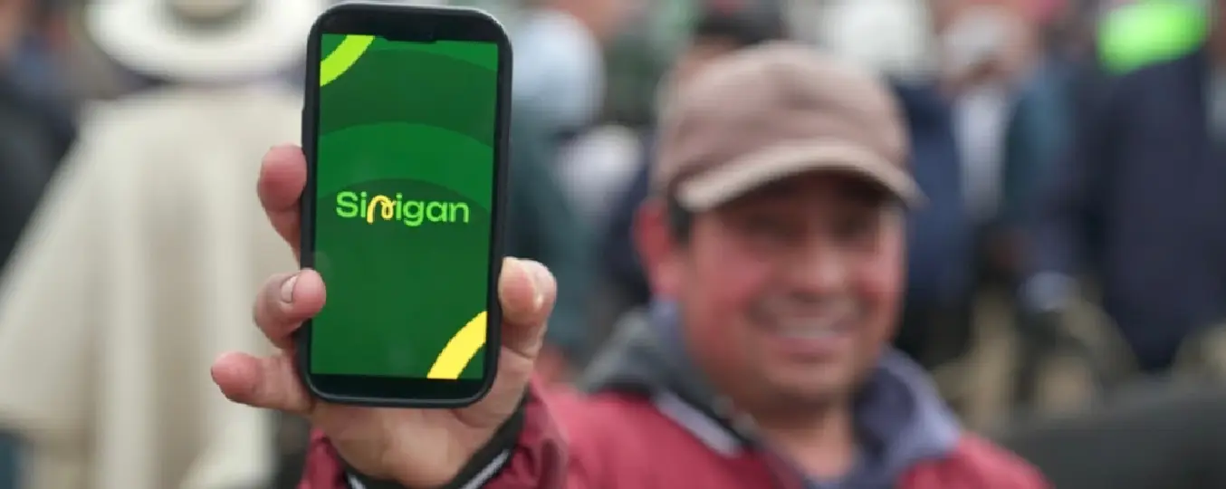A person in a red jacket and cap holding a smartphone displaying the Sinigan app screen, with a blurred crowd in the background.