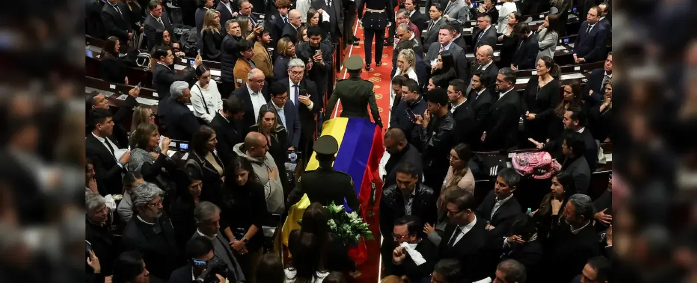 A coffin draped with a Colombian flag is carried through a crowded room of mourners dressed in black.