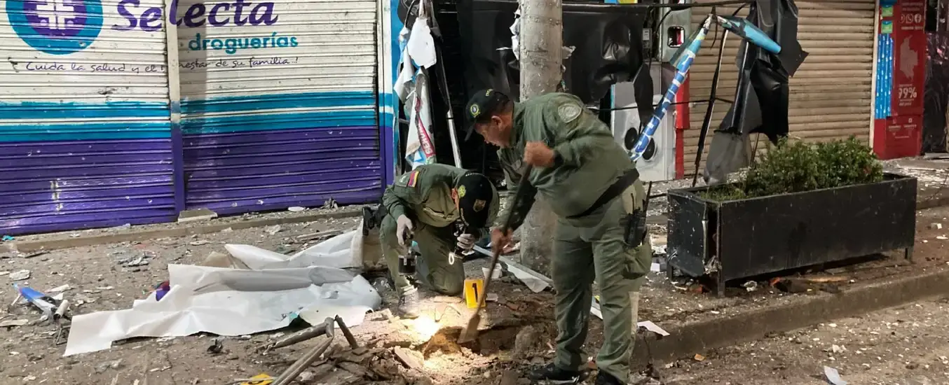 Two uniformed officers inspect debris on a street, using tools near a damaged storefront with scattered rubble and a partially collapsed awning.