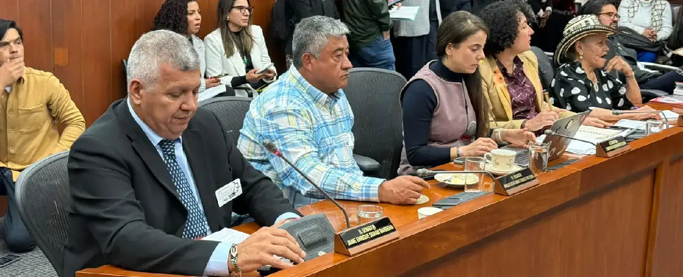 A group of people seated at a conference table, with microphones and nameplates, engaged in a discussion. Audience members are visible in the background.
