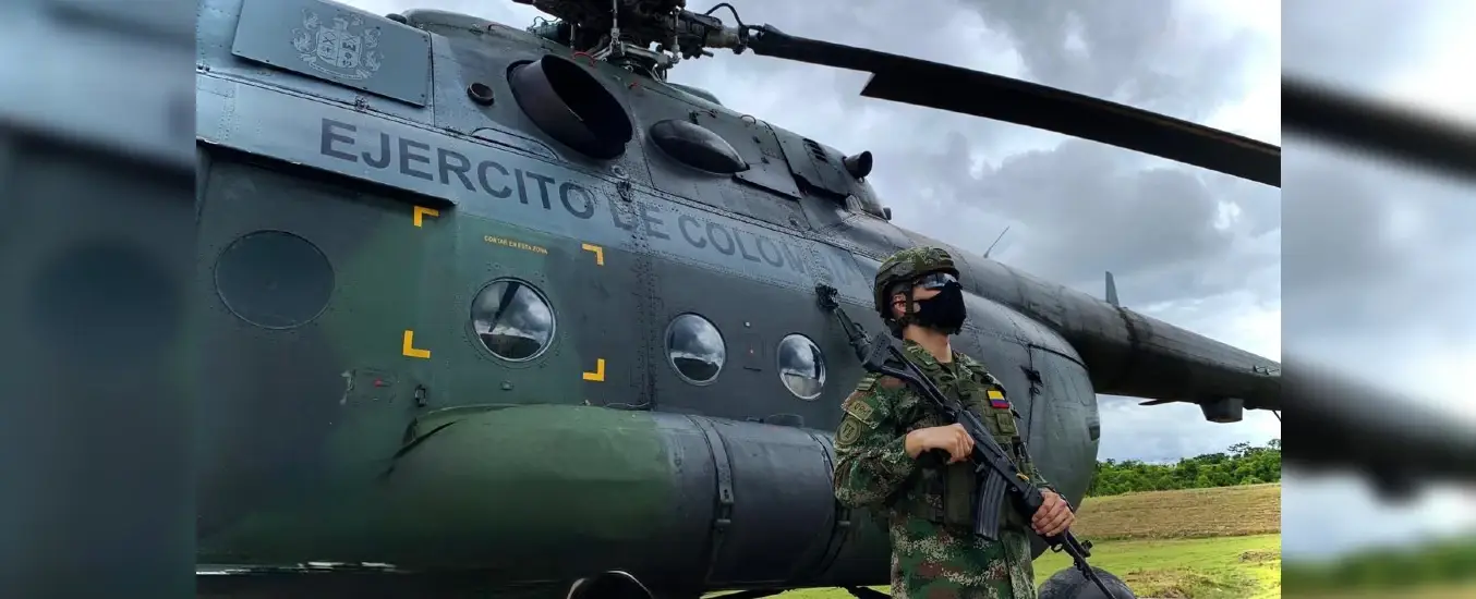 Soldier in camouflage and mask stands guard beside a military helicopter with "EJERCITO DE COLOMBIA" text, under a cloudy sky.