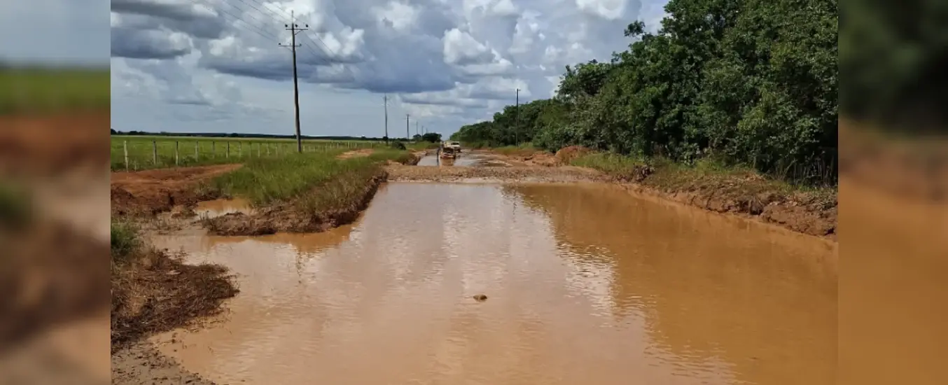 A muddy, waterlogged dirt road with a vehicle in the distance, surrounded by grassy fields and trees under a cloudy sky.