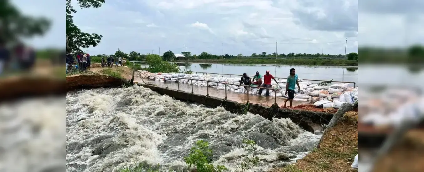 Floodwaters breach a barrier with sandbags, as people stand on a flooded road beside a turbulent river under a cloudy sky.