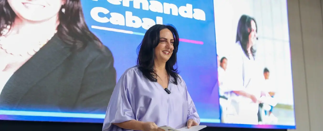 A woman in a light purple blouse stands smiling, holding papers, in front of a large screen displaying her image and name.