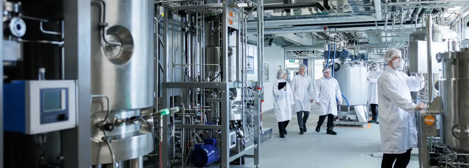 Científicos con batas blancas y máscaras caminan por un laboratorio industrial con grandes equipos de metal y paneles de control.