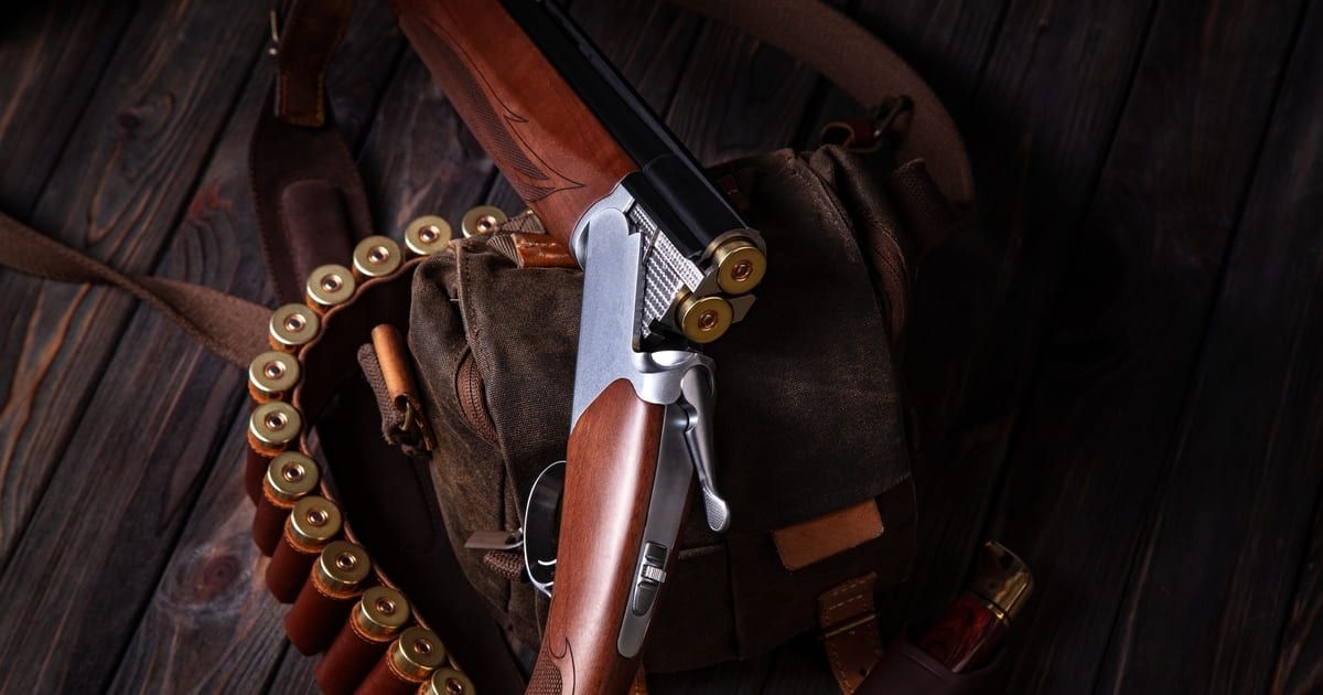 A shotgun with two bullets inside, laid on a rustic background with a brown bag and a strap of shotgun shells.